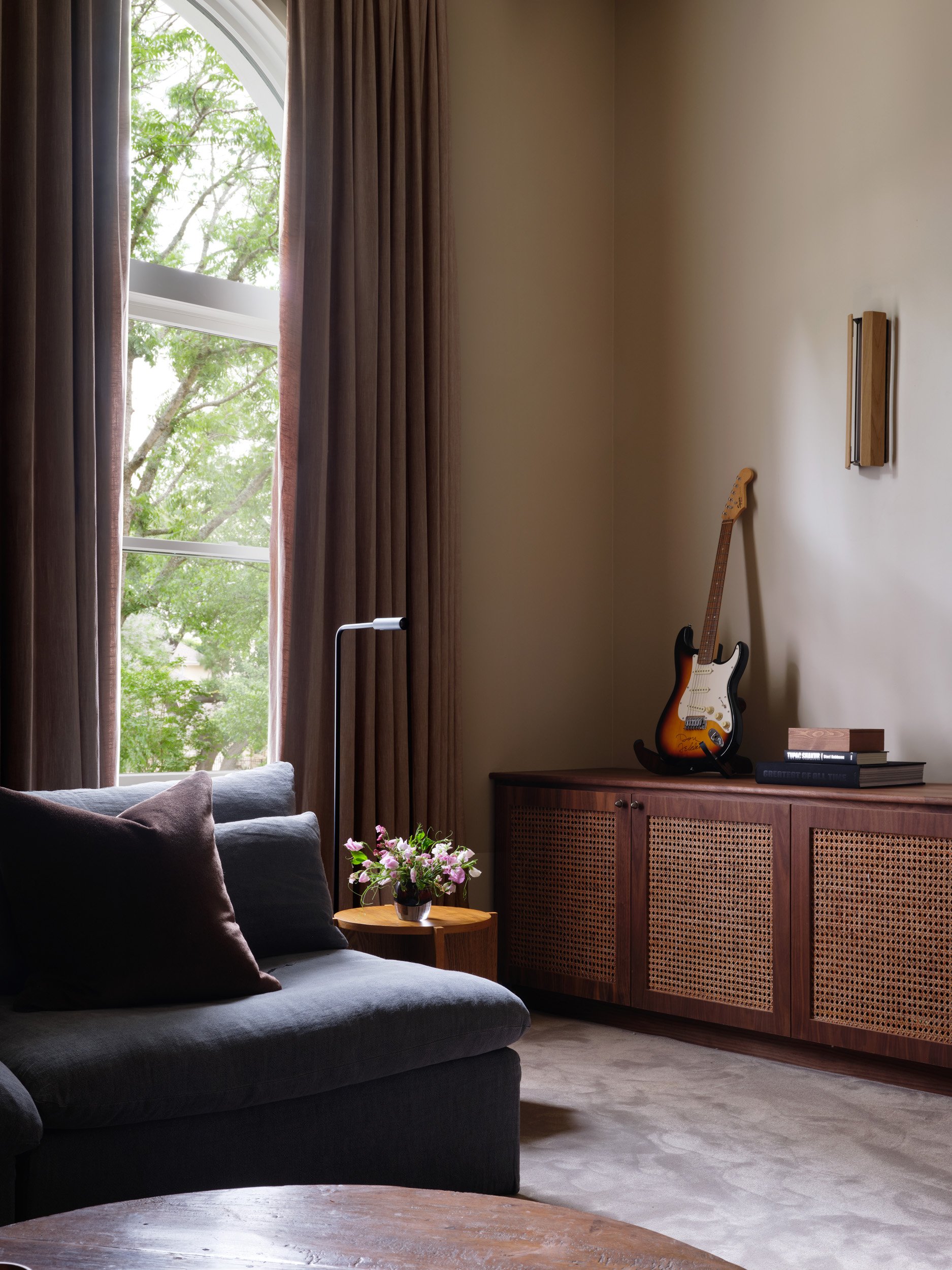 Living room corner with gray sofa, dark pillows, small wooden side table with flowers, large window with brown curtains, guitar on sideboard, books on sideboard.