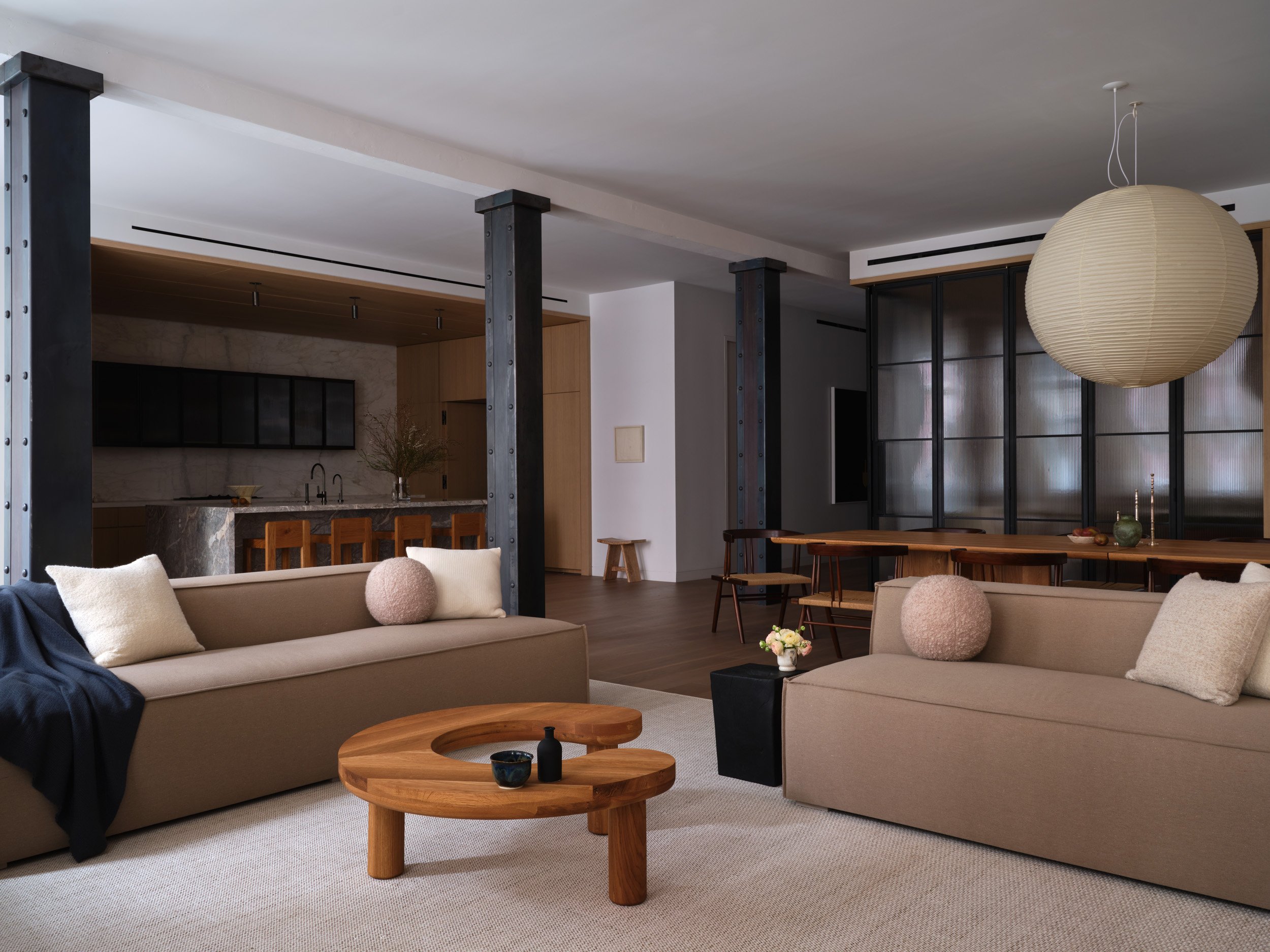 Modern open-concept living room and kitchen with beige sofas, wooden coffee table, black accents, and a large round paper lantern hanging from the ceiling.