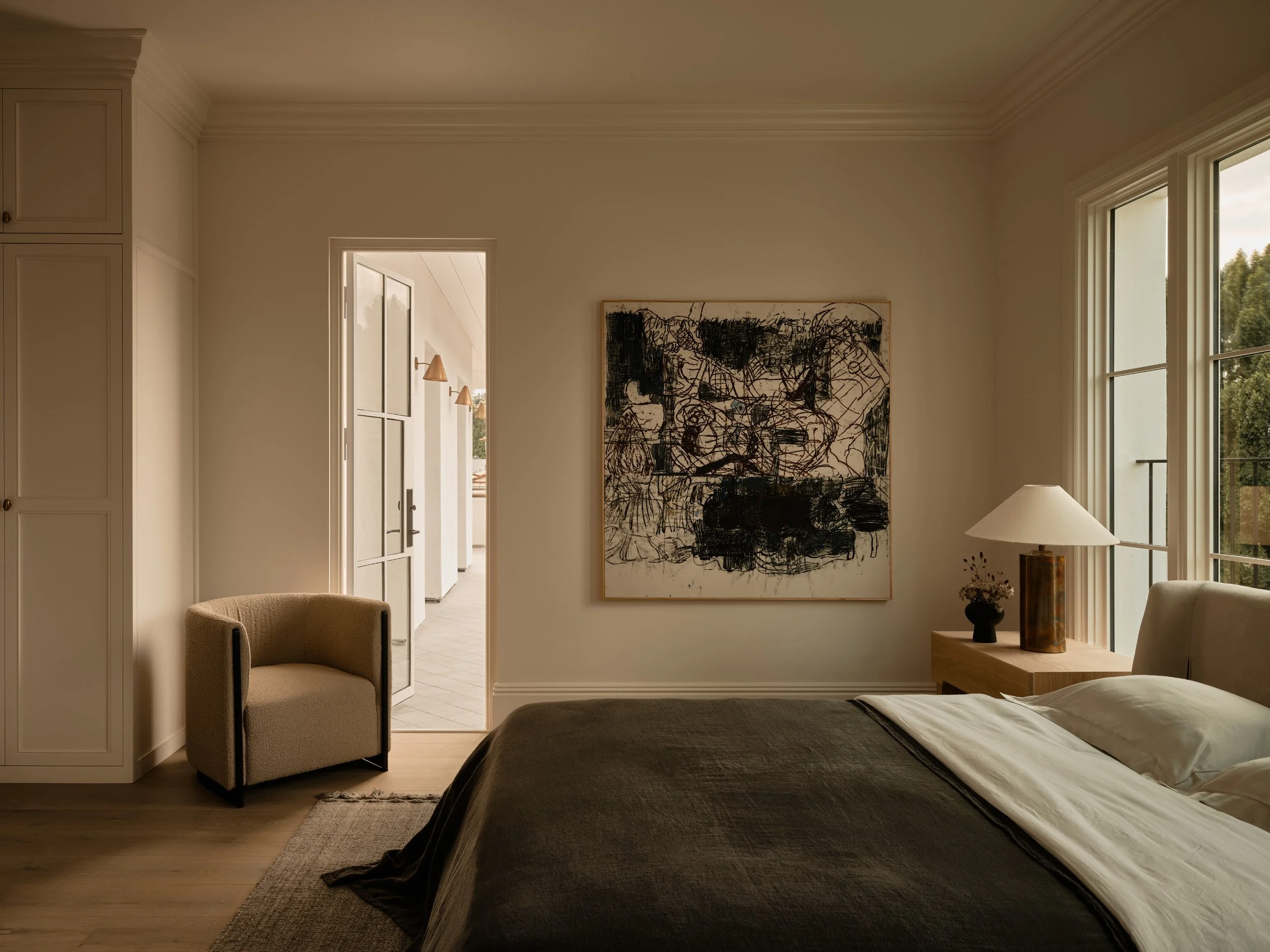 A bedroom with a bed, large window, abstract artwork on the wall, and a small armchair near the doorway, with a view into a hallway.