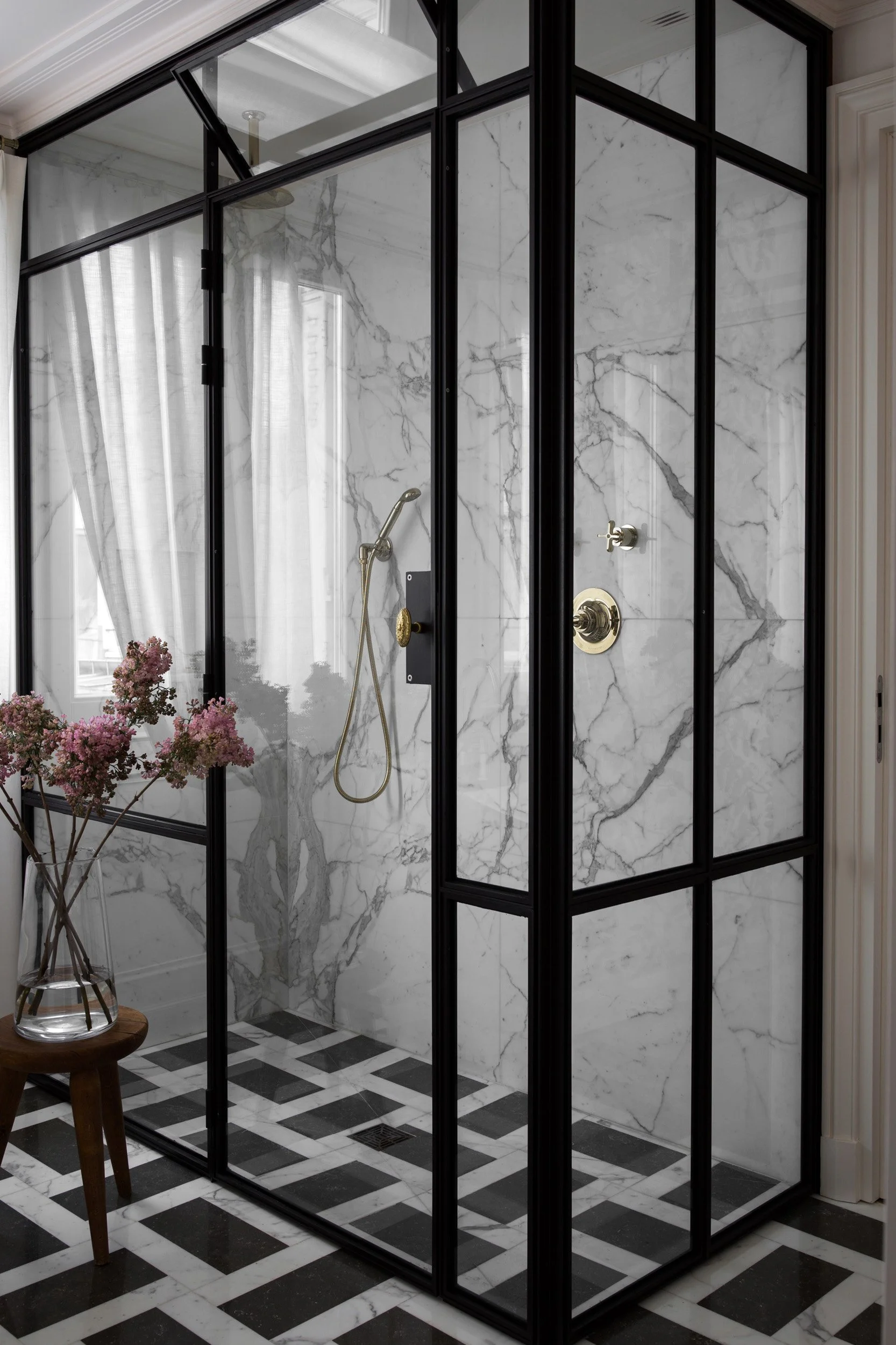 Modern walk-in glass shower with marble walls and black metal framing, featuring a brass showerhead and fixtures, with black and white checkered tile flooring and a small wooden stool with pink flowers.