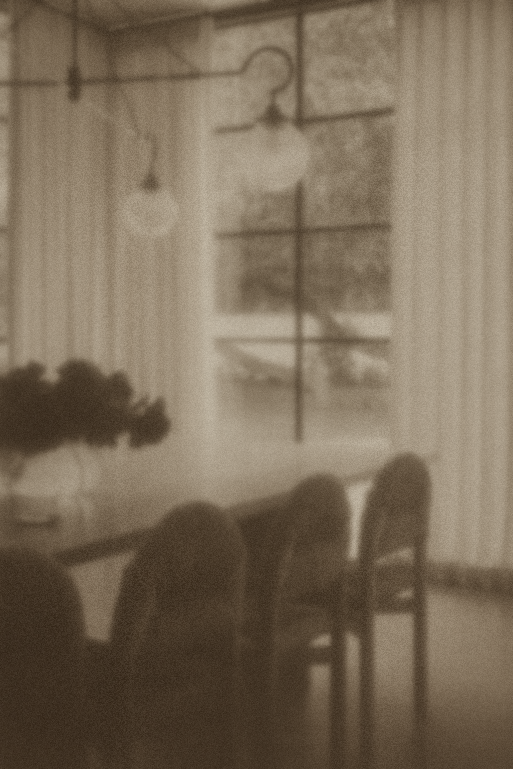 A sepia-toned image of a cozy dining area with a rectangular table, four chairs, a potted plant, and a window with a cross-style window pane. The room has wooden walls and hanging pendant lights.
