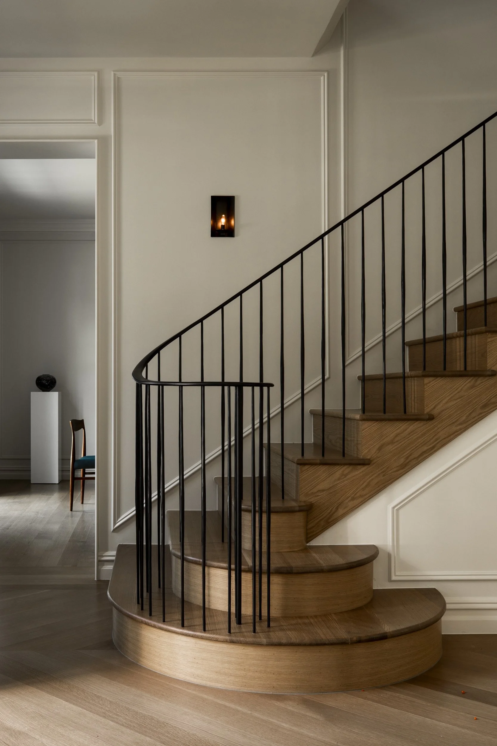 A modern indoor stairway with wooden steps and a black metal railing, located next to a white wall with decorative molding, and a small wall sconce with a candle inside.
