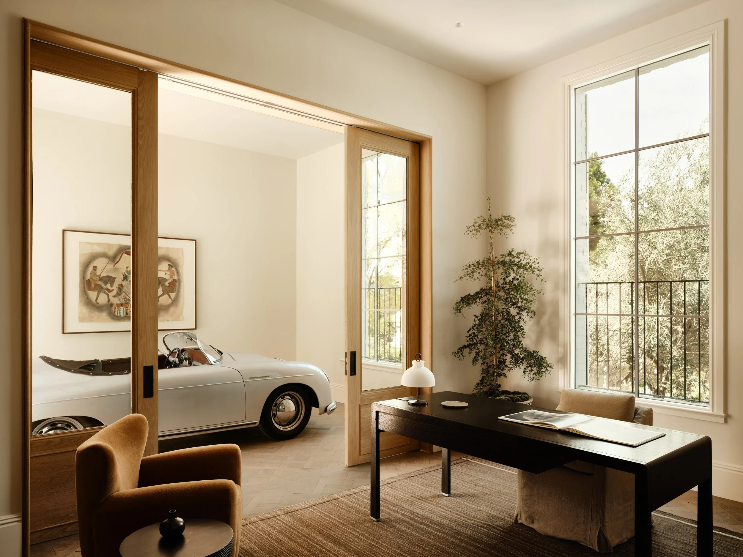 Sunlit room with large windows, a black desk with an open book and a small lamp, a potted tree, and a brown armchair, with a sliding door leading to a room containing a white vintage car and a framed artwork.
