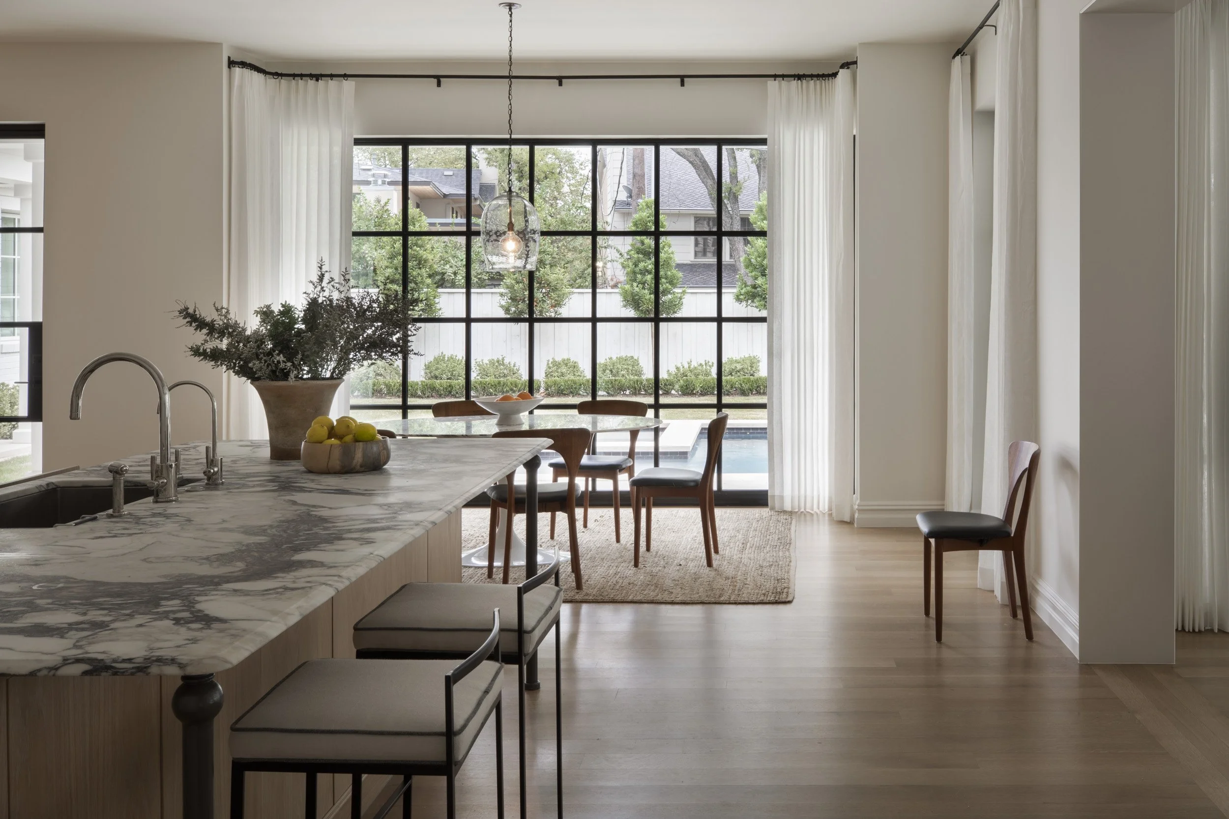 A modern dining area with a large window view of a backyard with trees and shrubs, featuring a marble kitchen island with a faucet, a bowl of lemons, a potted plant, a dining table with chairs, and white curtains.