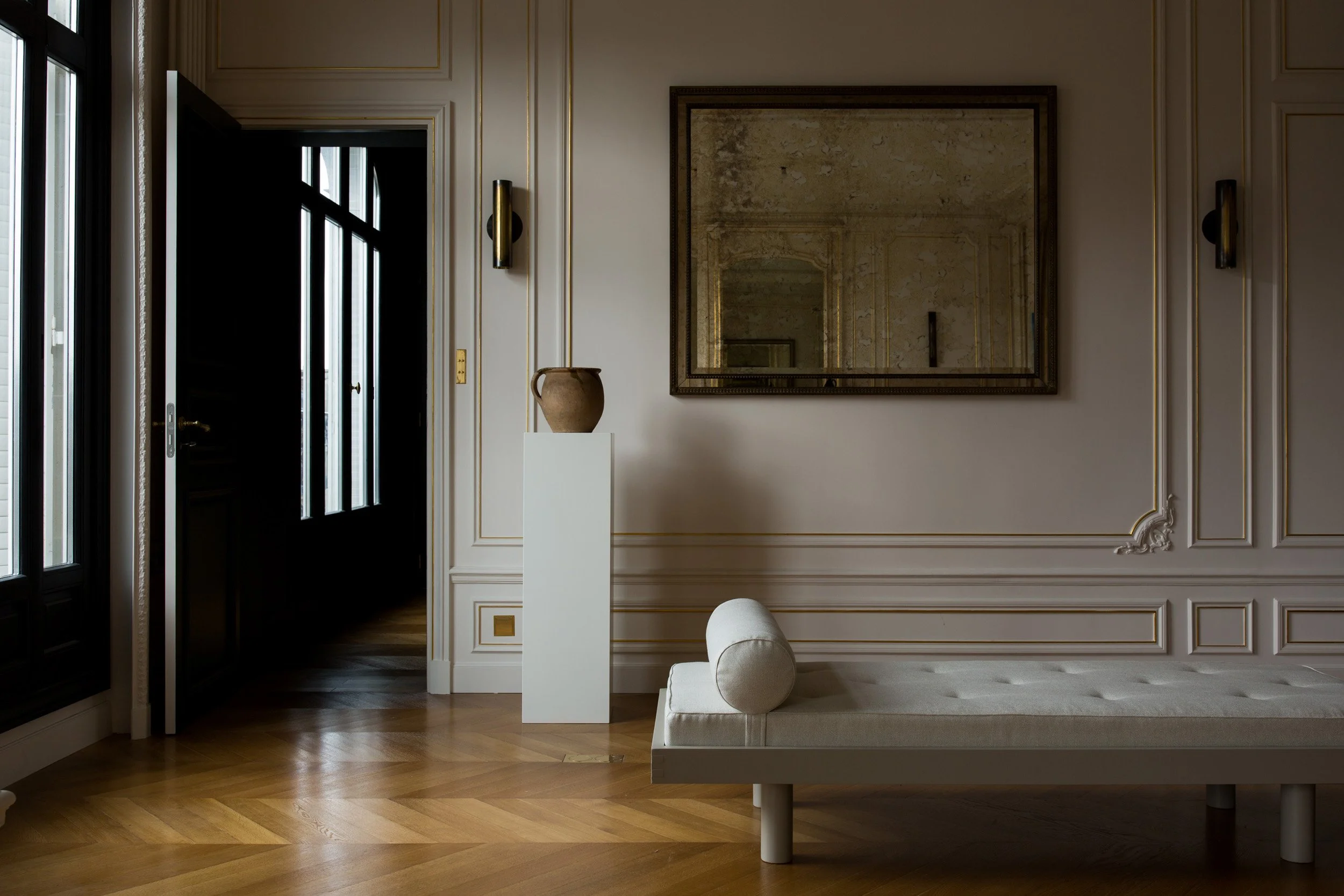 Minimalist interior with wooden flooring, white walls with decorative molding, a mirror, a bench with a cushion, a tall white pedestal with a clay pot, and a black door with glass panels.