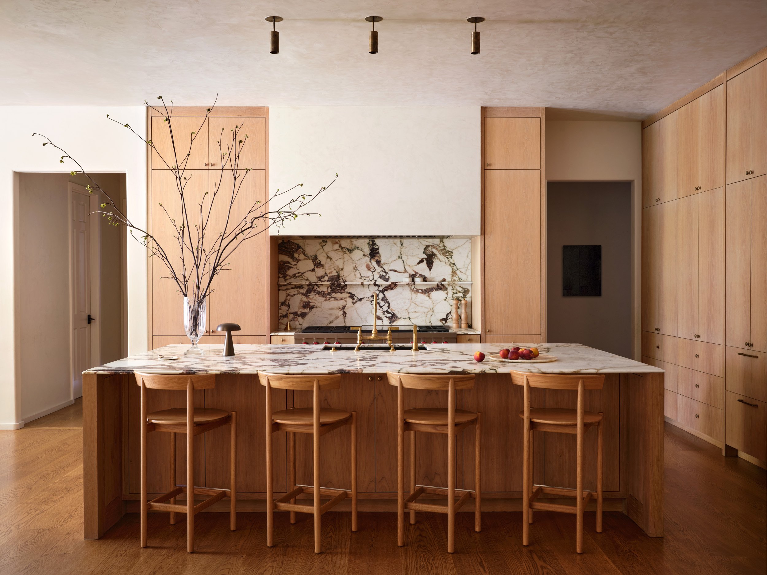 Modern kitchen with a large marble island, wooden cabinetry, and a marble backsplash, decorated with a tall vase of branches and a small plate of peaches.