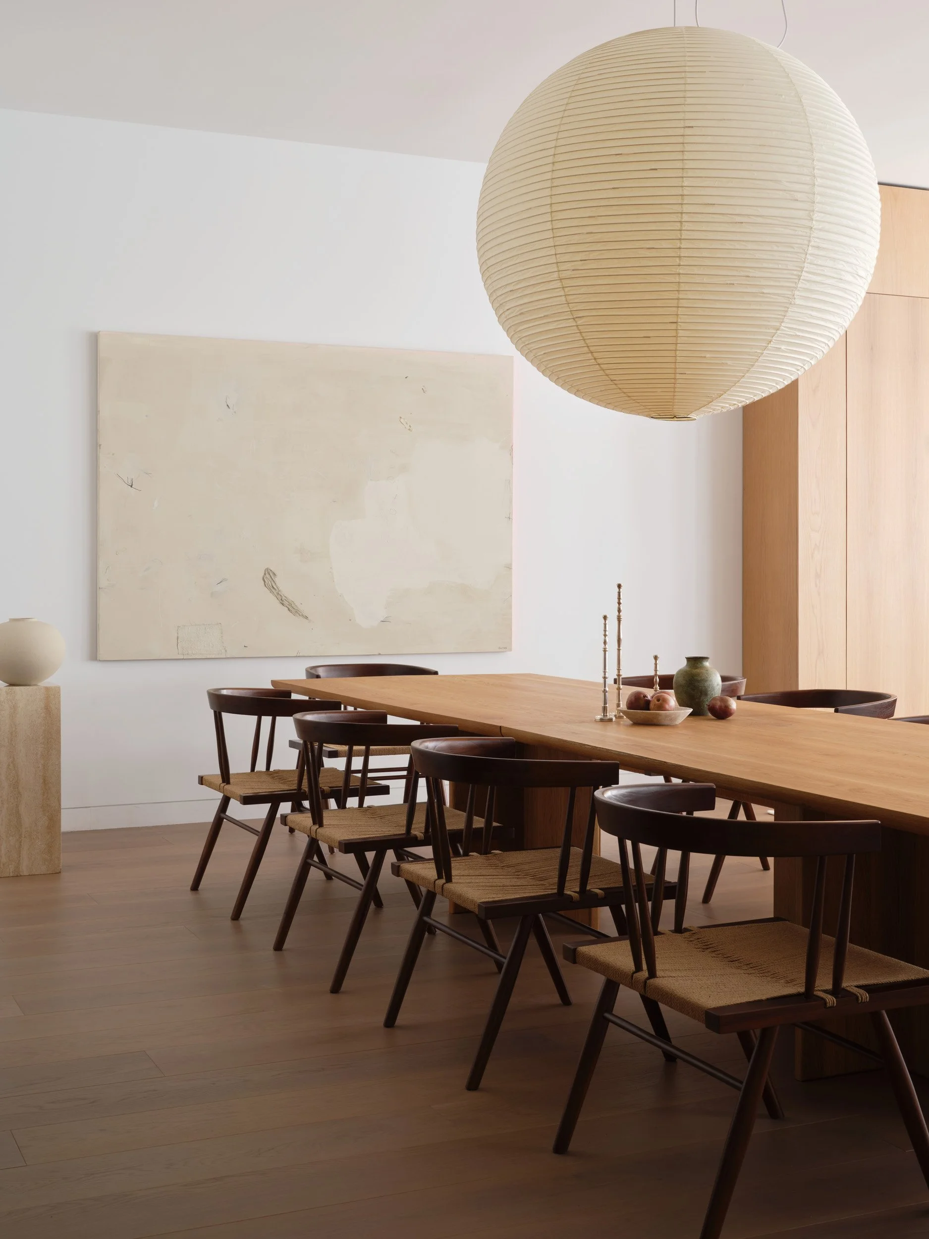 A minimalist dining room featuring a large wooden table surrounded by six dark wooden chairs with woven seats. A large paper lantern hangs overhead, and there is an abstract painting on the white wall behind the table. The floor is hardwood, and there are simple decor items, including vases and candlesticks, on the table and a nearby pedestal.
