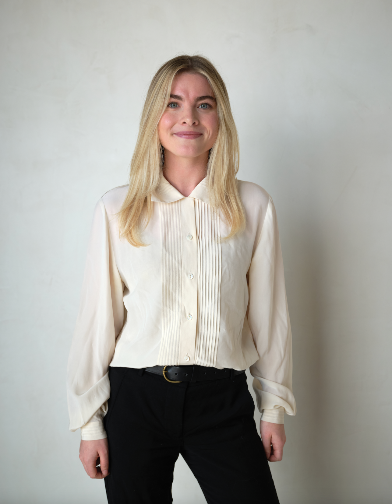 A woman with blonde hair in a cream blouse and black pants standing against a plain white background.