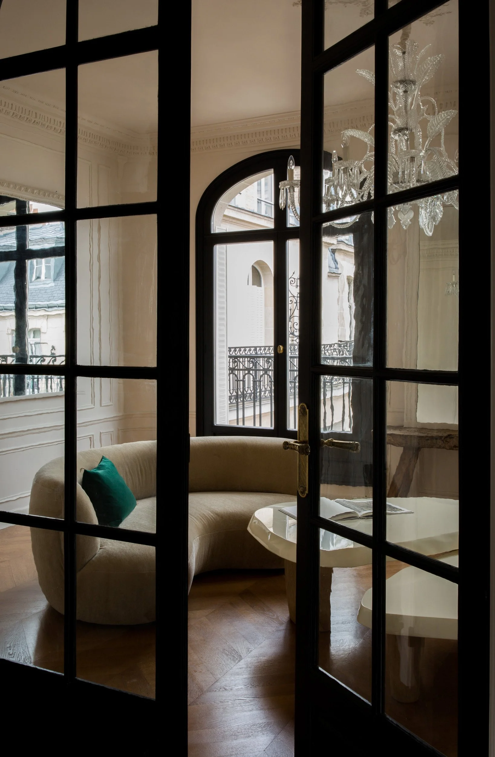 Interior view of a cozy living room with a beige curved sofa, a small round marble coffee table with a book, large glass doors opening to a balcony, and a crystal chandelier hanging from the ceiling.
