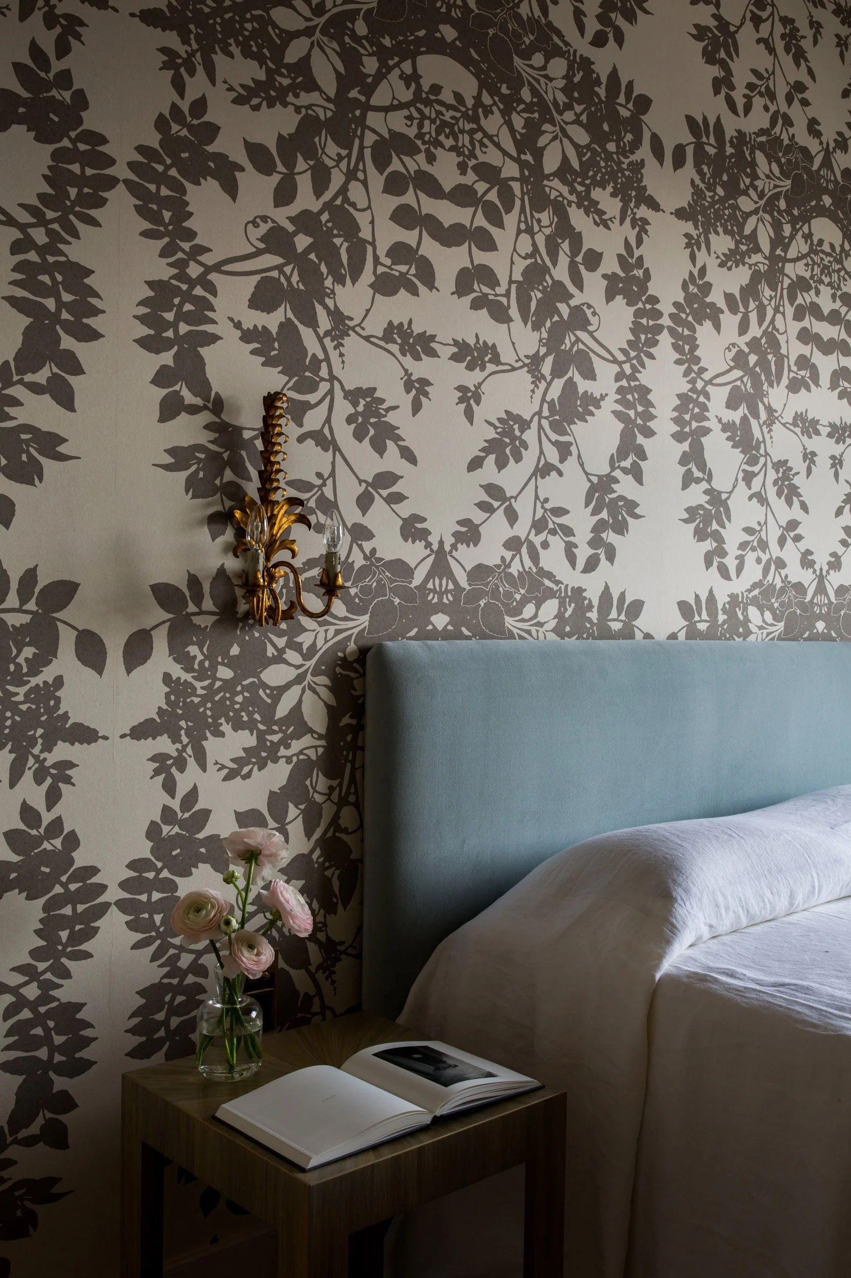 A bedroom with a wallpaper of dark leaf and branch patterns, a brass wall sconce with light bulbs, a light blue upholstered headboard, a wooden nightstand with an open book, a clear vase with pink and white flowers, and black-and-white photographs on the book.