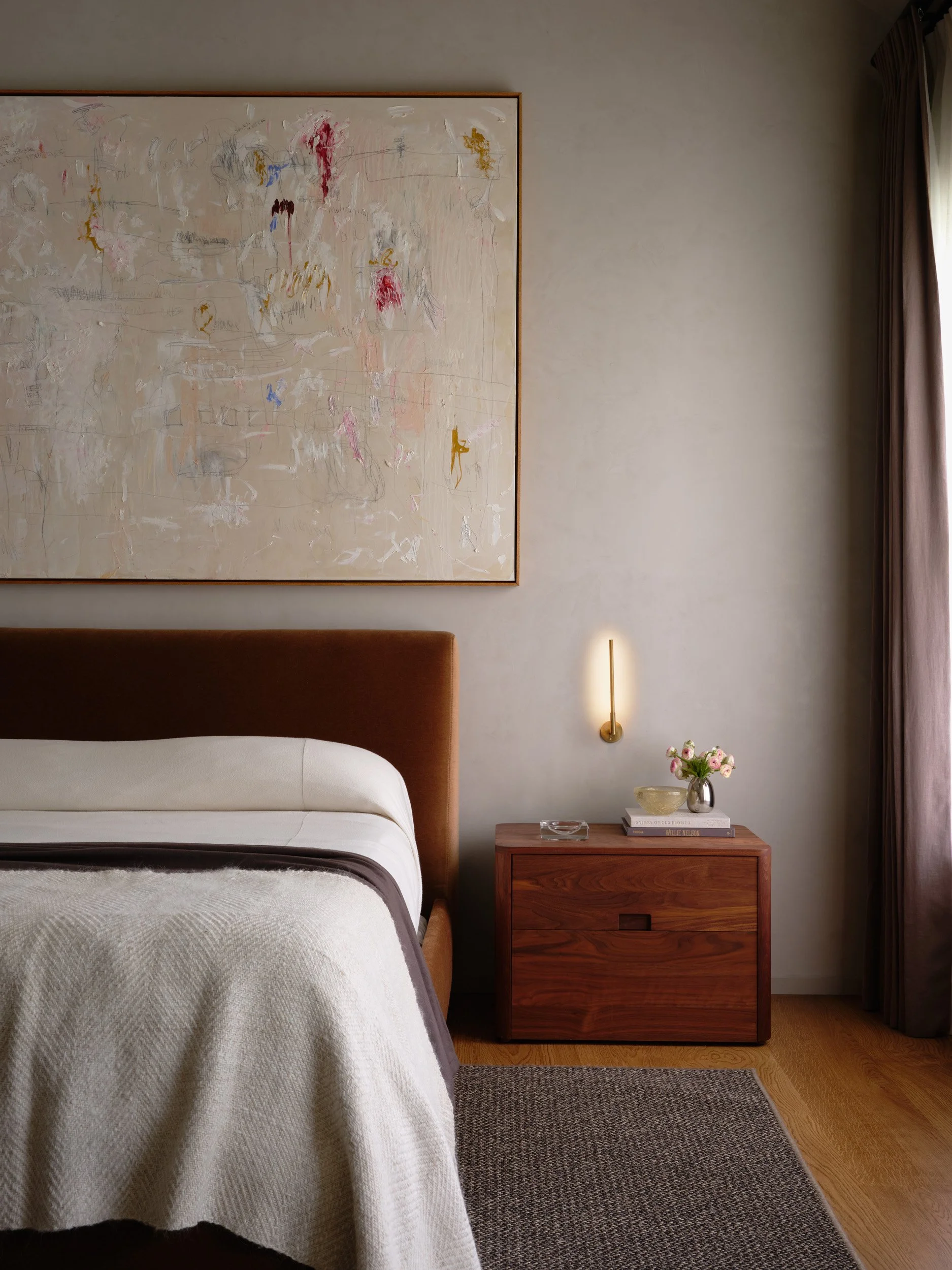 A bedroom with a bed, an abstract painting above, a wooden nightstand with a vase of flowers, a lamp, and some books, near a window with curtains.
