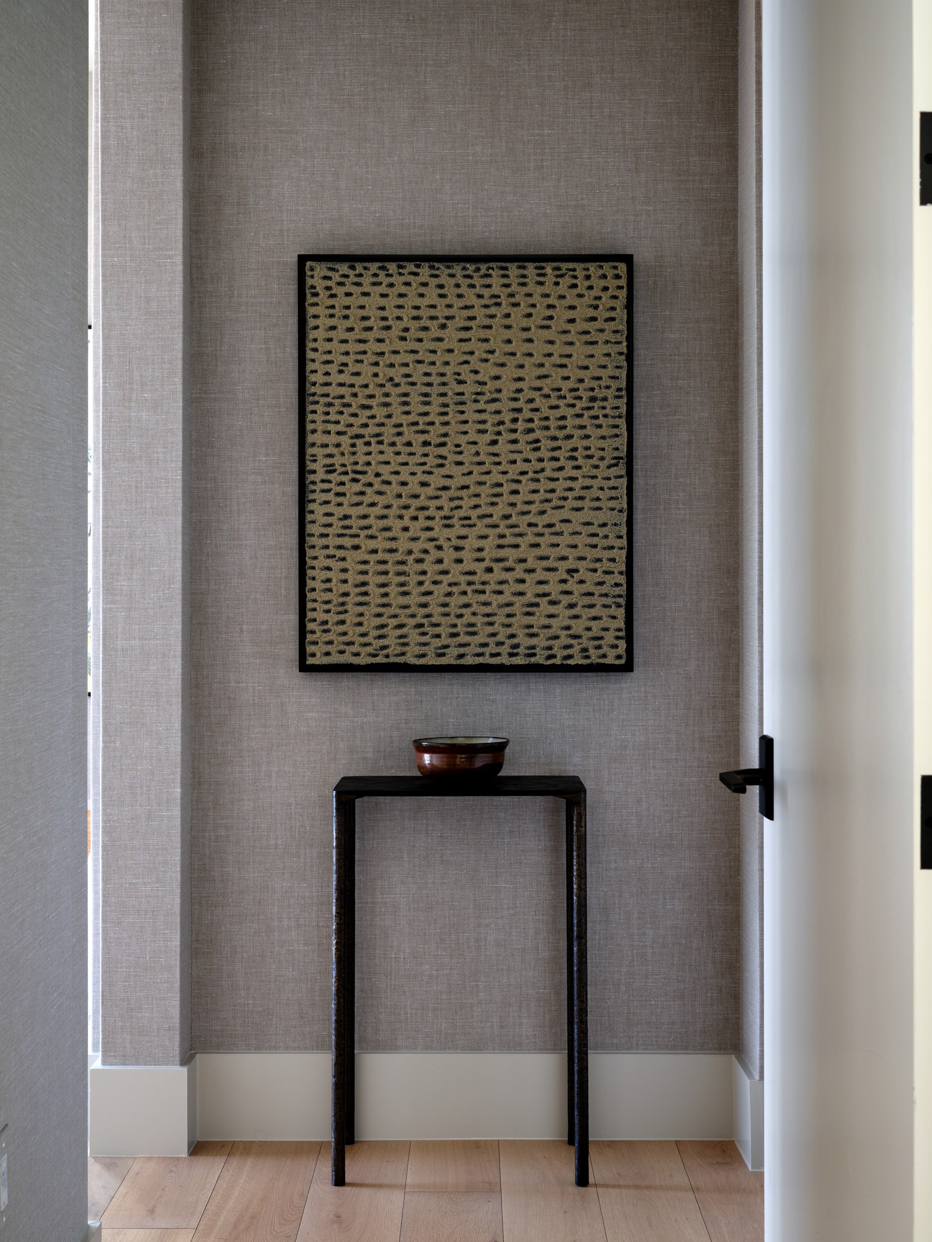 A small wooden table with a bowl on top, set against a wall with textured beige wallpaper, decorated with a framed piece of abstract art.
