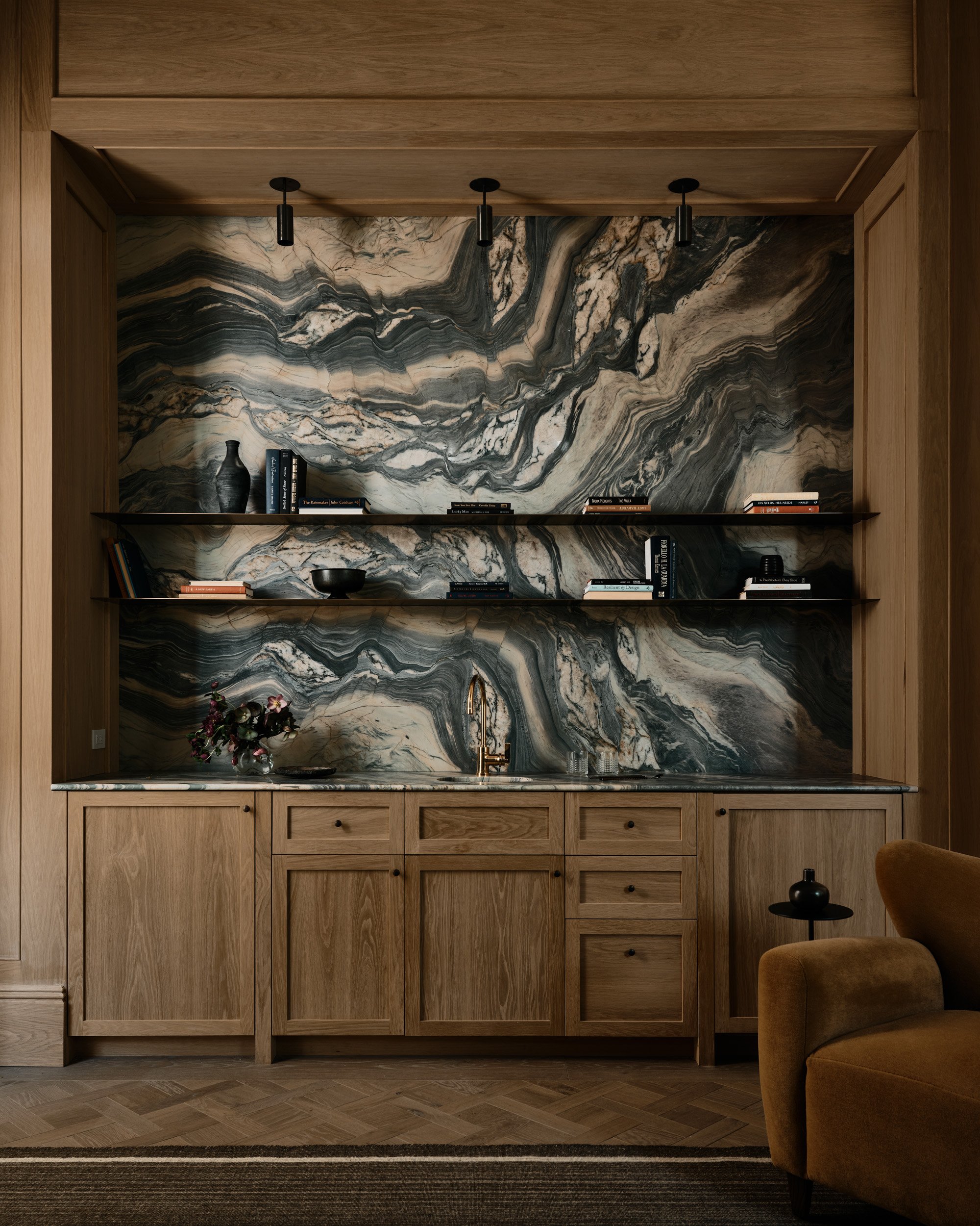 A wooden kitchen cabinet with a marble backsplash, open shelving with books and decorative items, and a small counter with a flower arrangement and a faucet.