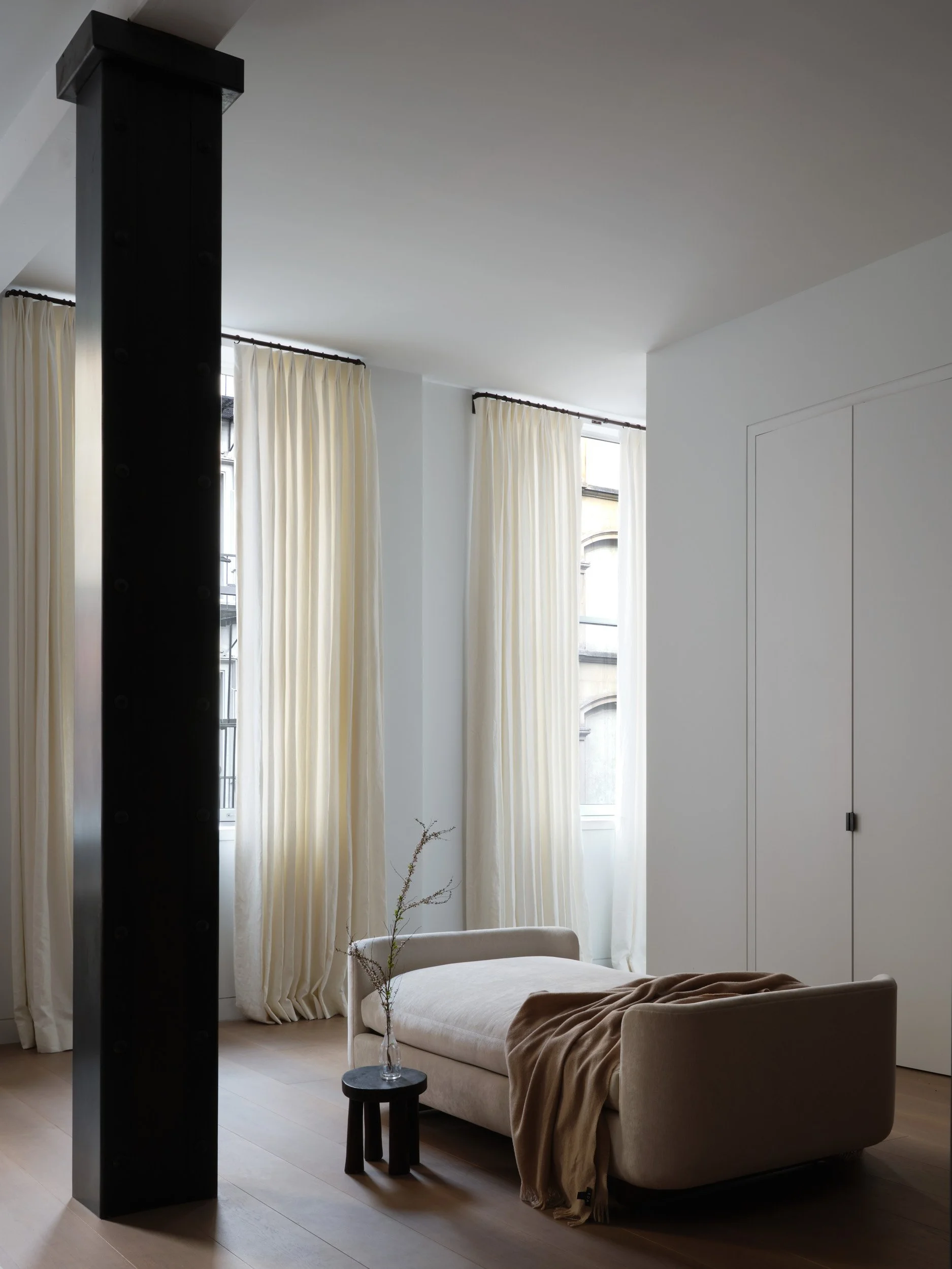 Living room with large windows, cream-colored curtains, a beige chaise lounge with a brown blanket, a small black side table with a glass vase holding a plant, and a black structural column.