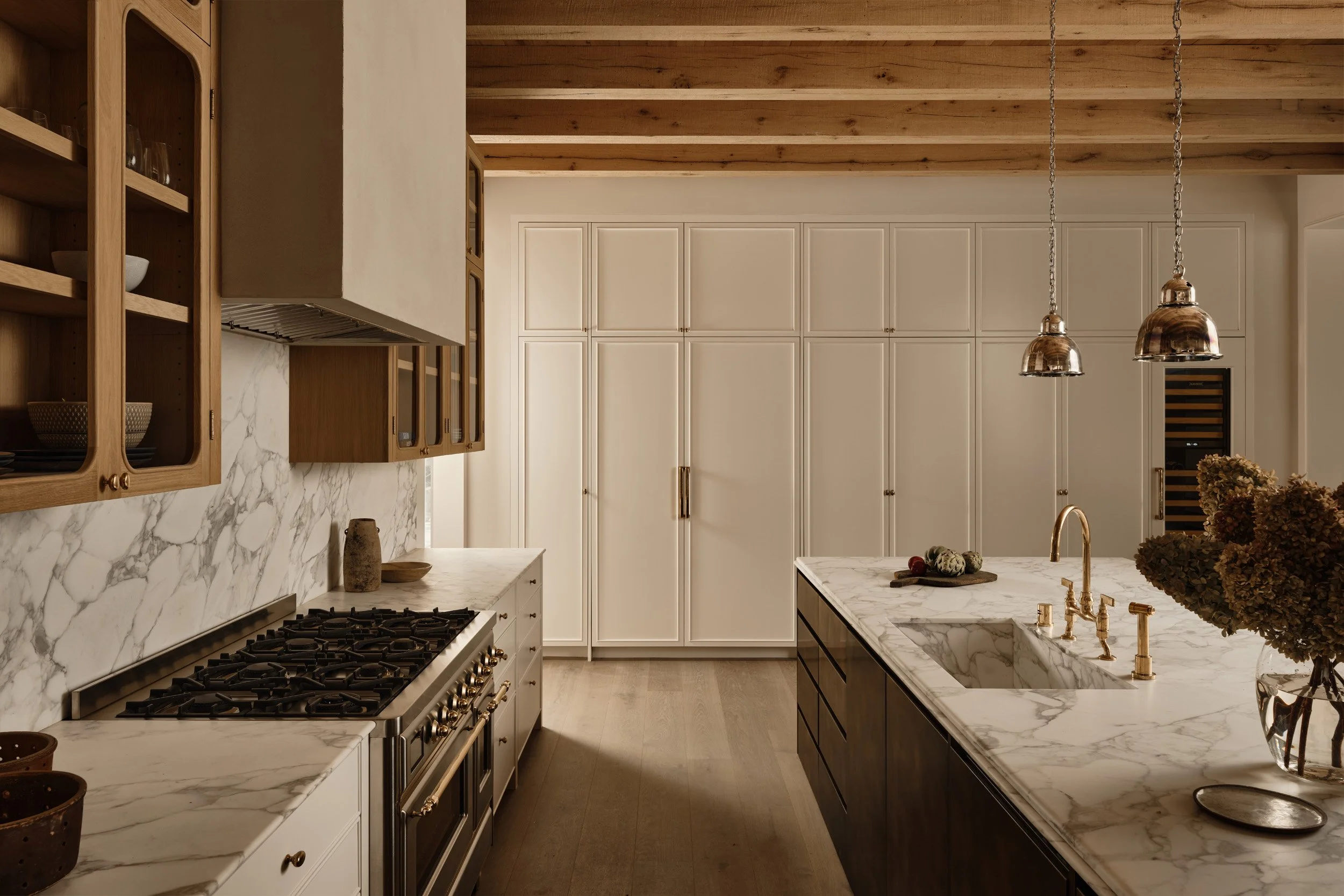 Modern kitchen featuring white marble countertops and backsplash, a large island with a sink, brass fixtures, wooden open shelves, and built-in white cabinets on the wall. Pendant lights hang over the island.