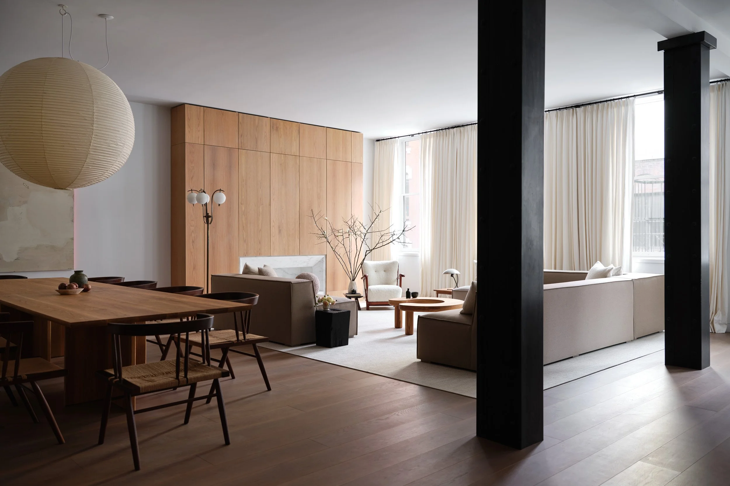 Living room with beige couches, wooden table, large windows with white curtains, tree branches in a vase, and a wooden wall panel.