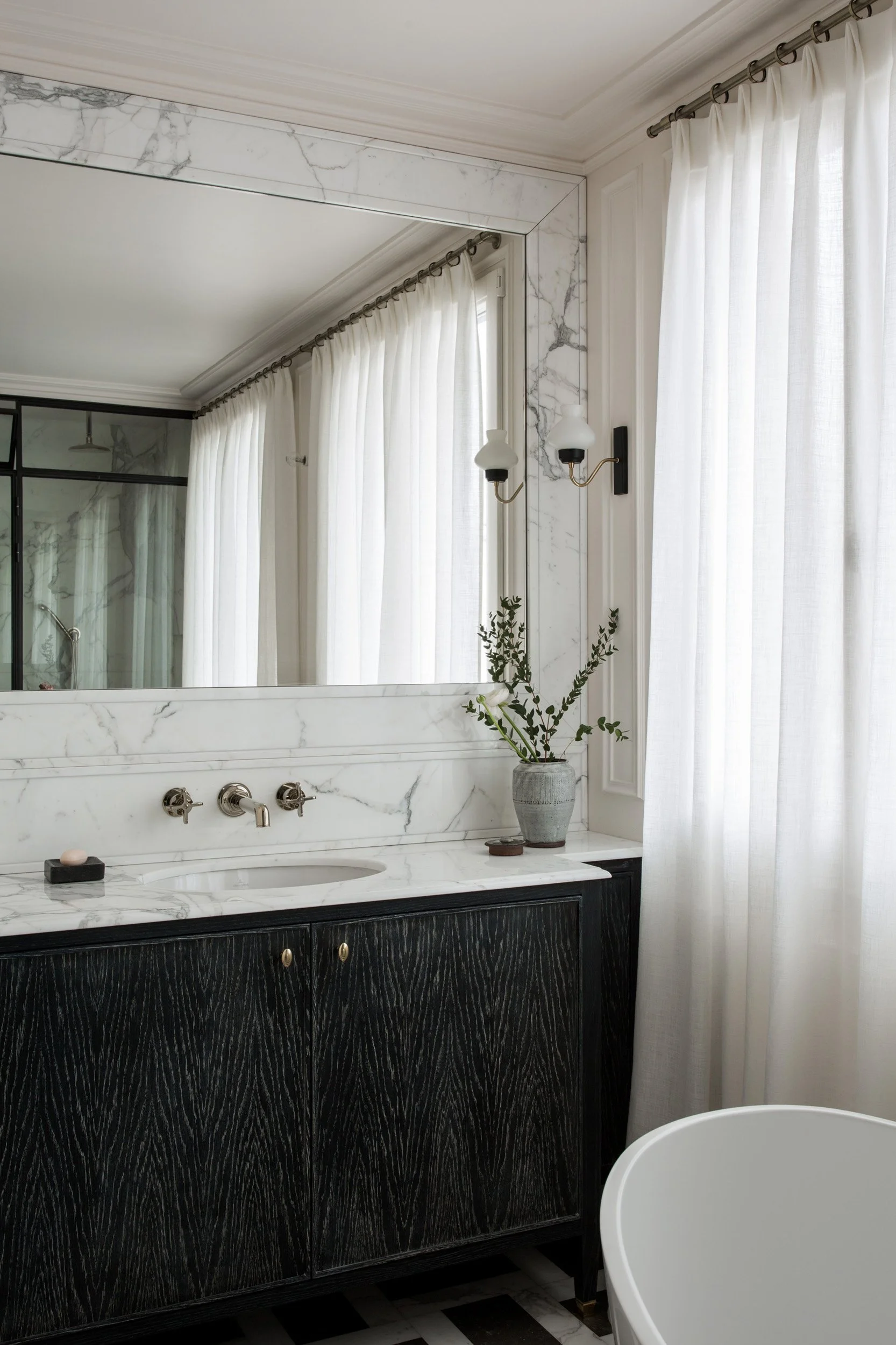A modern bathroom with a white marble countertop and backsplash, black wooden cabinet, large mirror, white curtains, and a sink with vintage-style silver fixtures. There is a vase with greenery on the counter and a bathtub partially visible in the foreground.