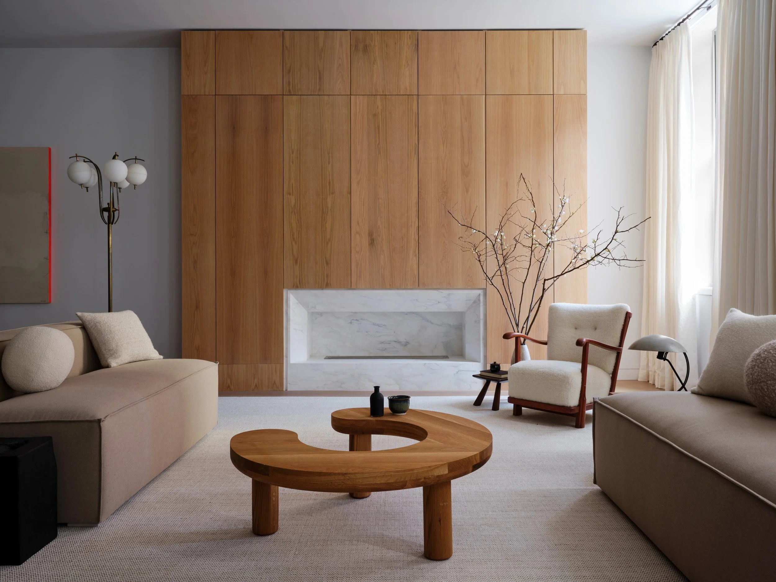 Living room with beige sofas, a wooden coffee table, a white armchair with wooden arms, a side table with a lamp, a tall floor lamp with three globes, a marble fireplace, a large window with cream curtains, a wooden wall panel, and a decorative branch with white blossoms.