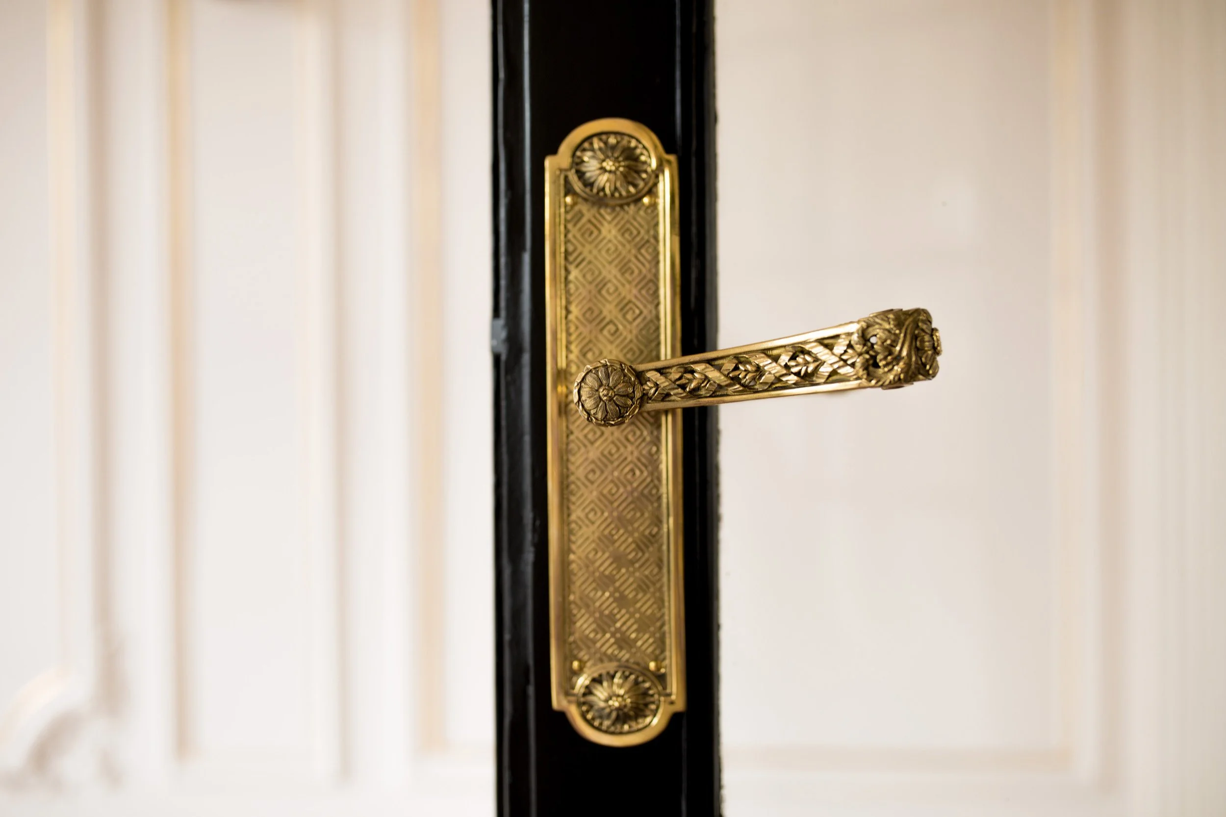 Close-up of an ornate gold door handle on a black door, with a cream wall in the background.