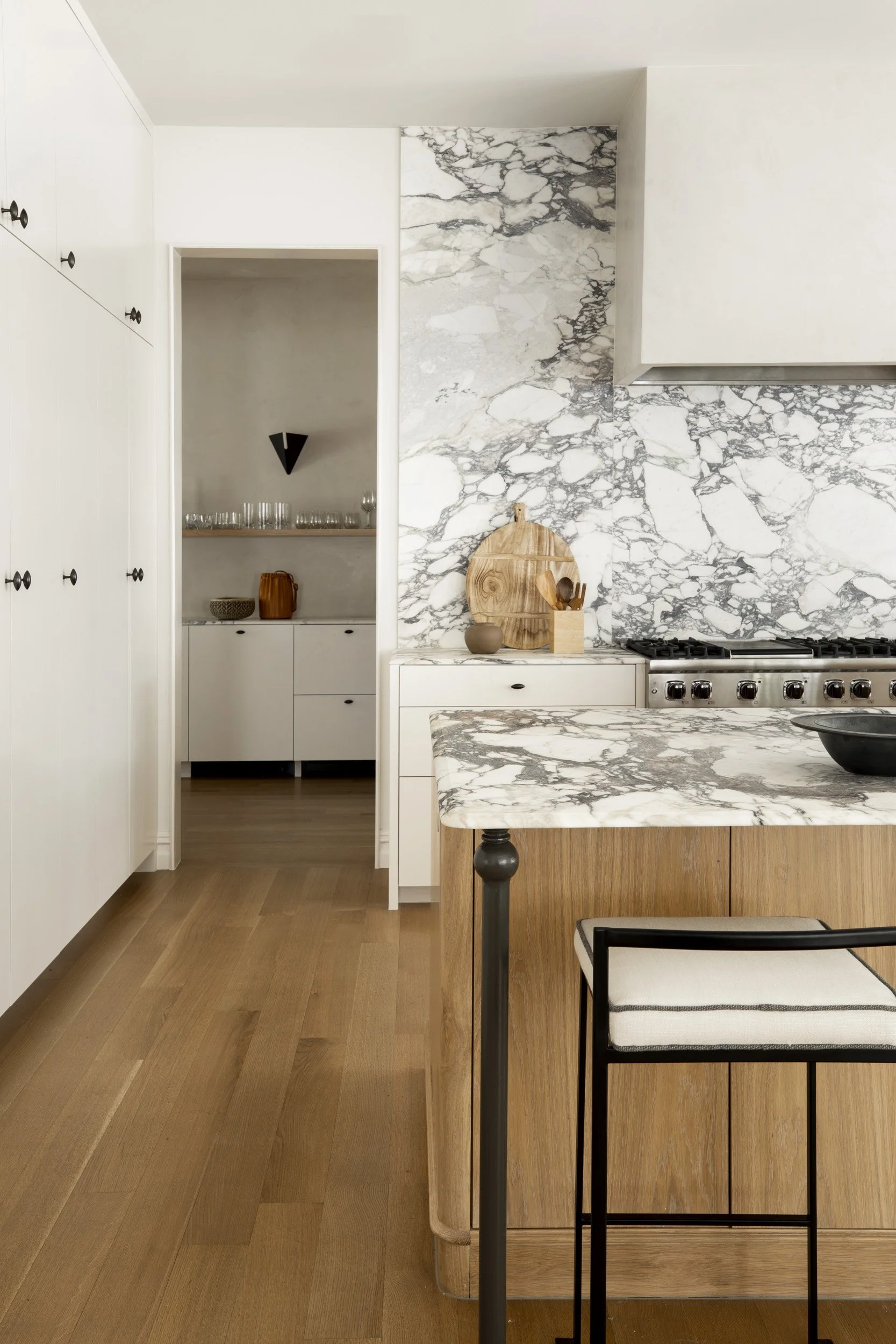 Modern kitchen with white cabinetry, marble backsplash, marble countertop, and wooden accents. There is a black and white stool in the foreground.