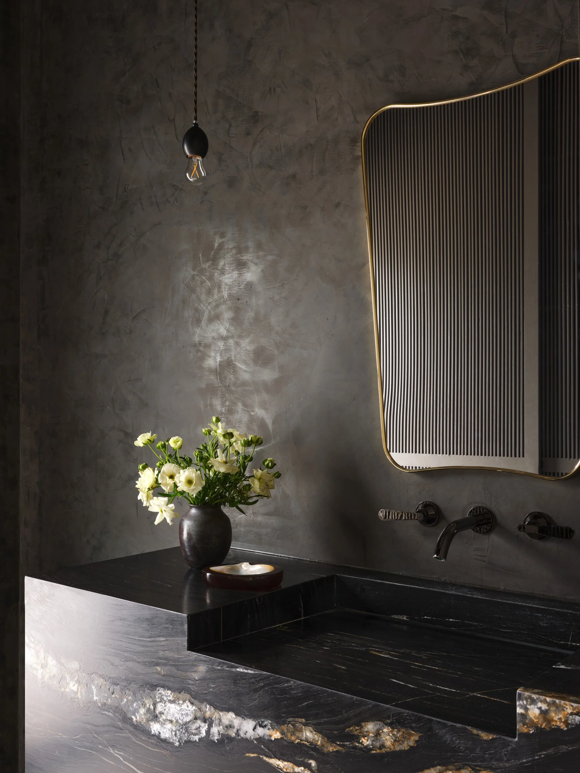 Modern bathroom sink area with a black marble countertop, a dark vase with white flowers, a small dish, a mirror with a gold edge, and a bare lightbulb hanging from the ceiling under a textured gray wall.