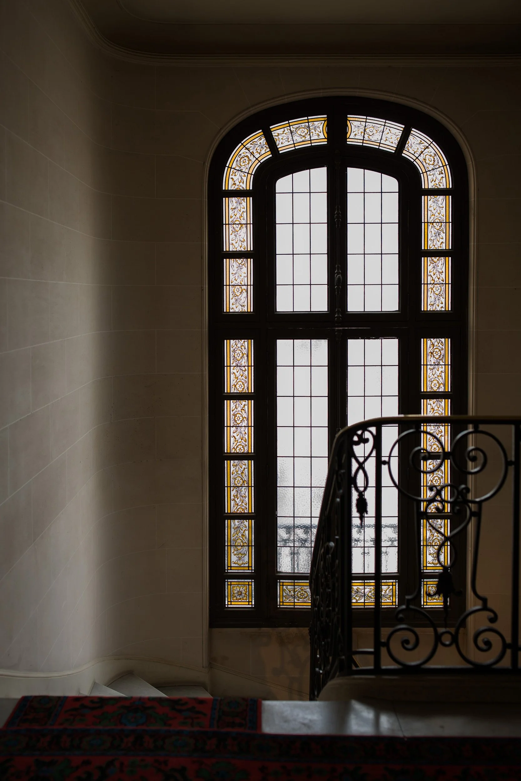 A large, arched window with stained glass accents and a decorative railing in front of it, inside a building with cream-colored walls.