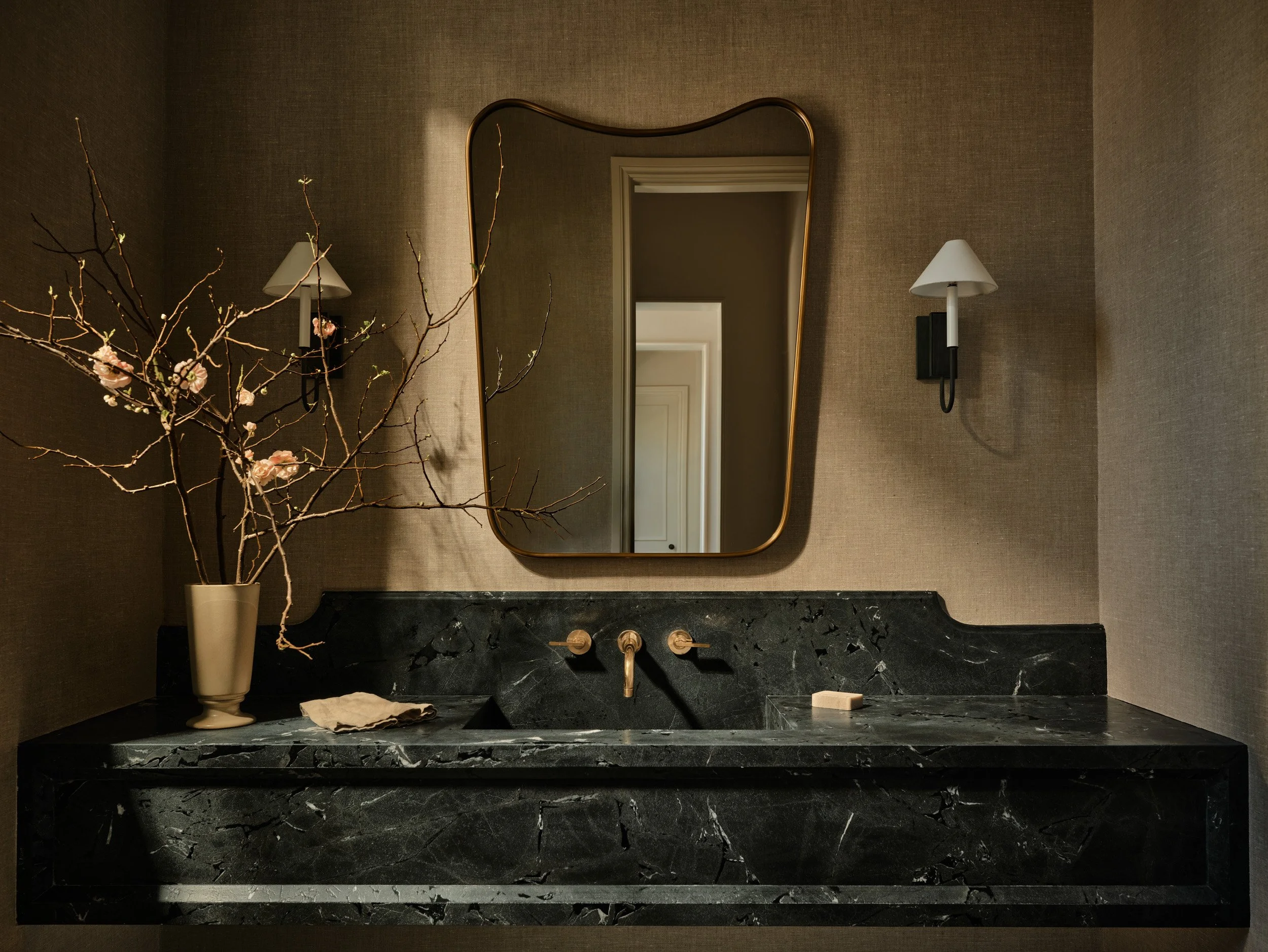 A bathroom vanity with a black marble countertop, a mirror with a gold frame, a vase with pink flowers and branches, a soap bar, and two wall-mounted lamps with white shades.