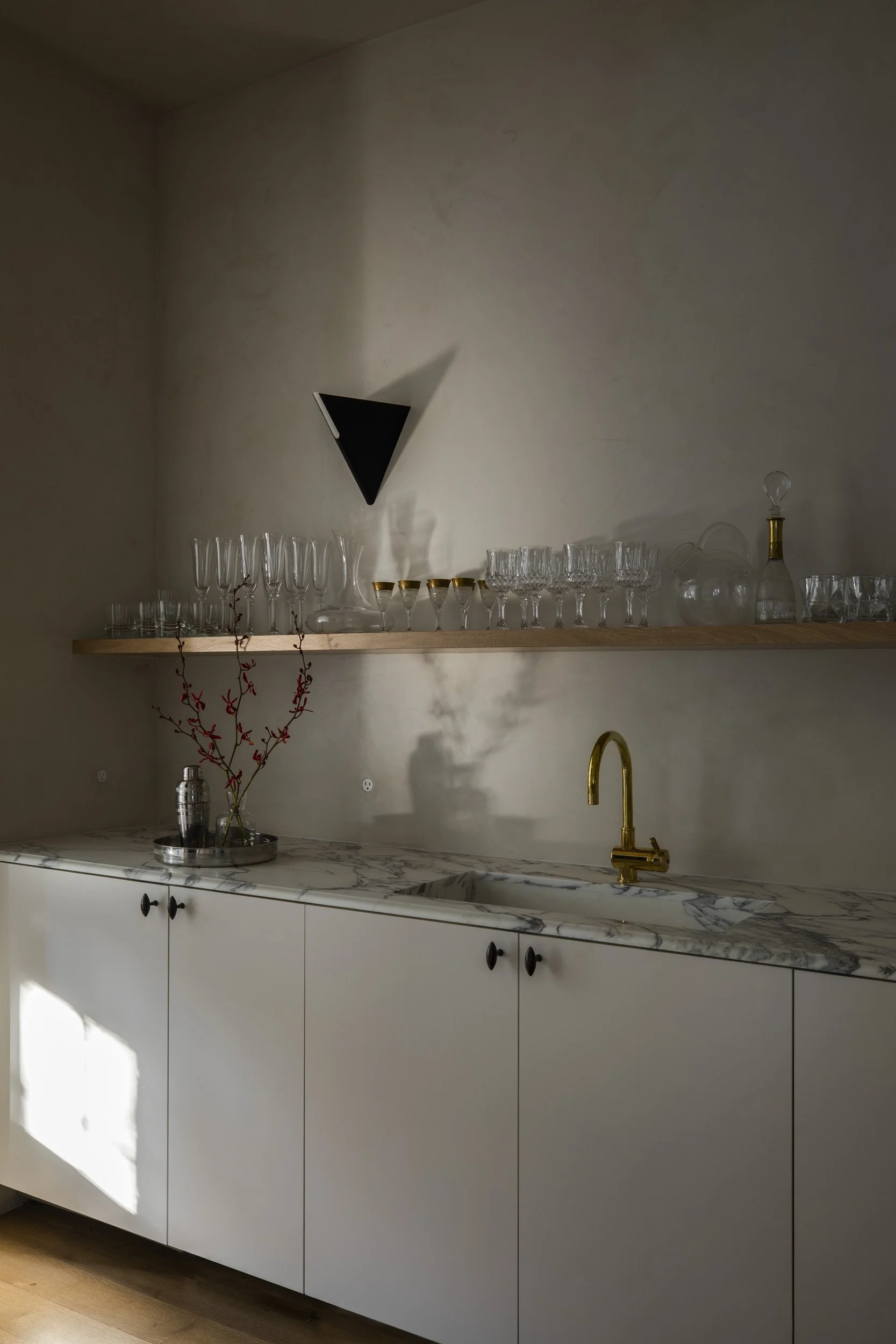 A minimalist kitchen with white cabinets, a marble countertop, a gold faucet, a wooden shelf with glassware, a vase with red branches, and shadows casting on the wall.