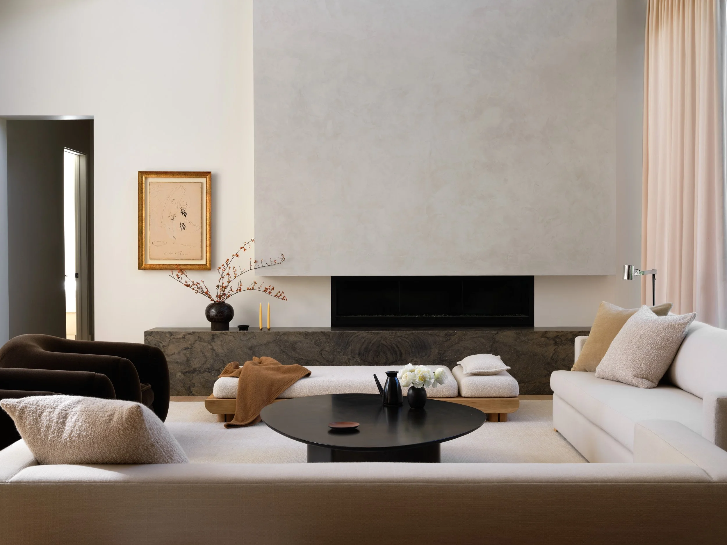 Modern living room with white sofa, black coffee table, stone fireplace, beige armchair, and decorative items including candles, vase with branches, and flowers.