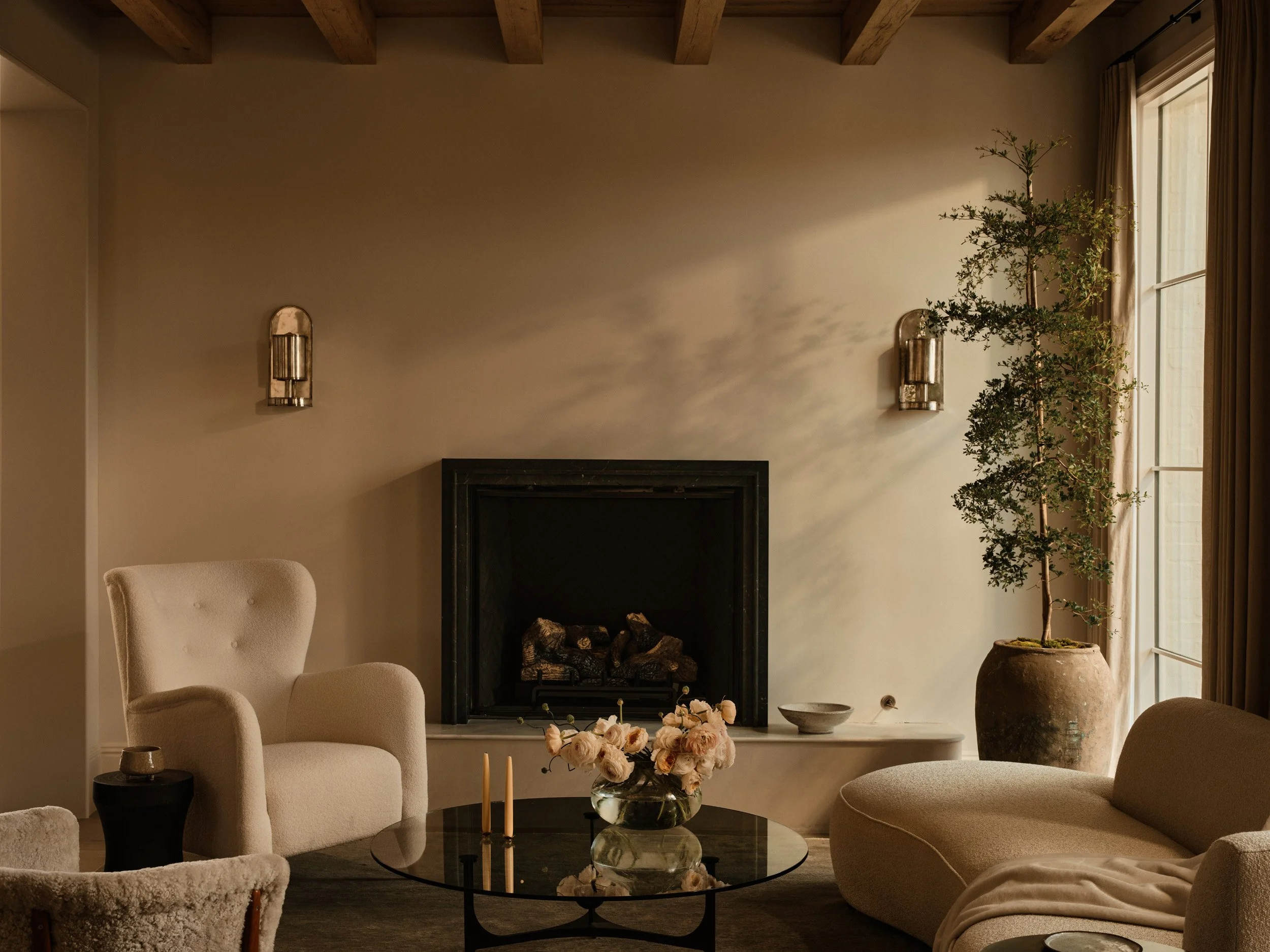 A cozy living room with a beige armchair, a sofa, and a glass coffee table with flowers. A fireplace with logs is centered on the wall, flanked by two wall-mounted lights. A large potted tree is near a window with curtains, and the ceiling has exposed wooden beams. Soft, warm lighting creates a calming ambiance.