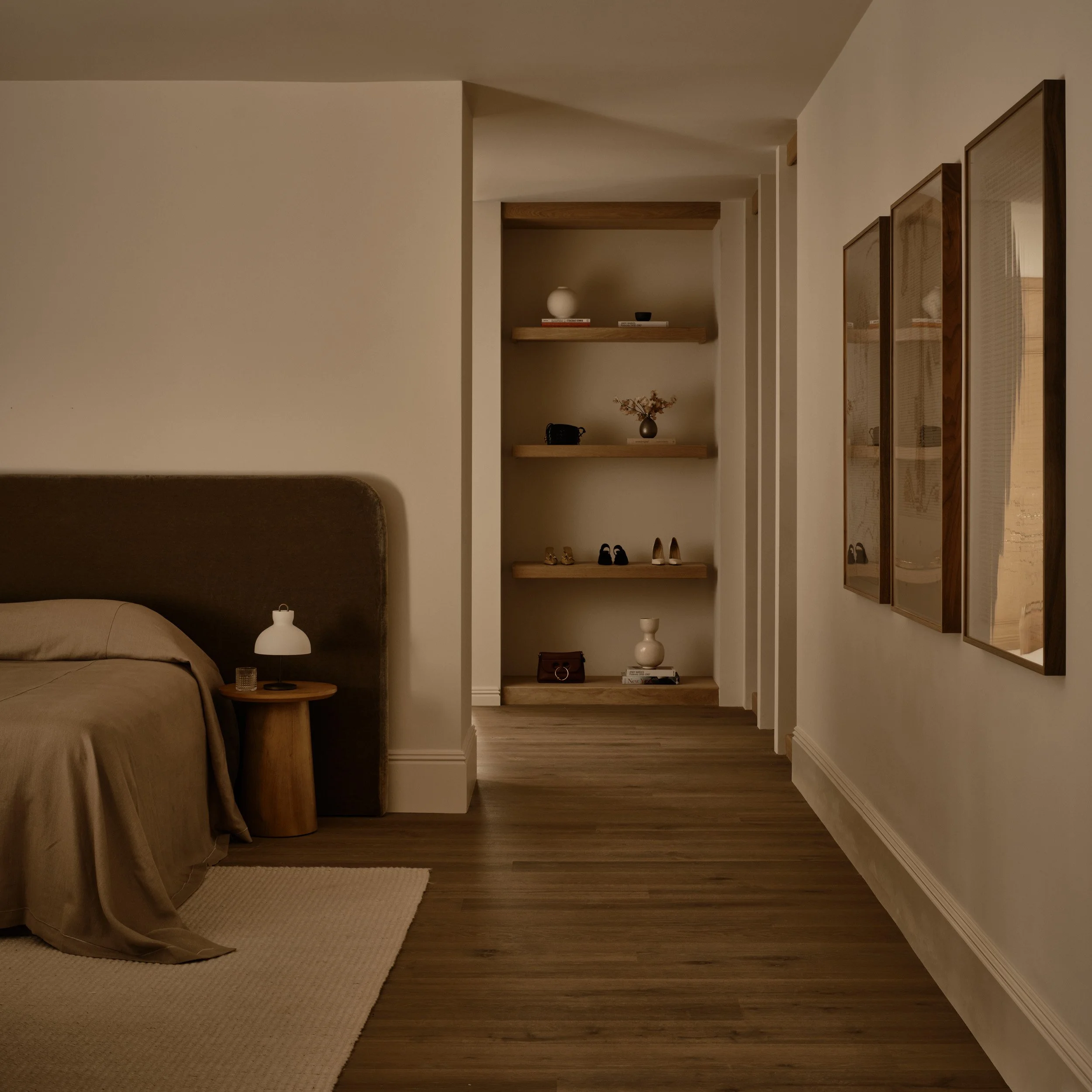 A bedroom with a bed, a nightstand with a lamp, and built-in shelves with decorative items. The room has a wood floor and framed art on the wall.