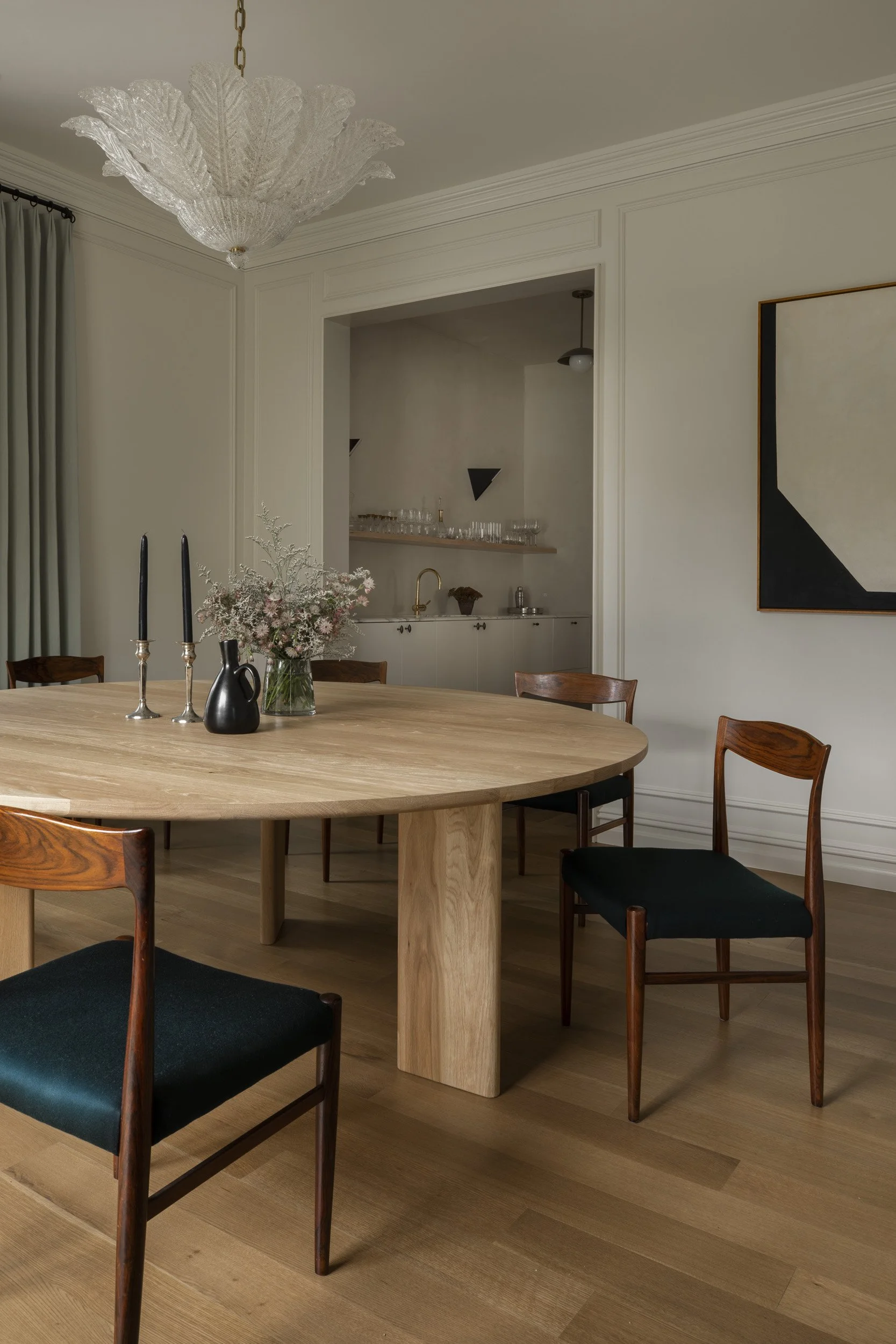 Modern dining room with wooden round table, black cushioned chairs, a chandelier, abstract wall art, and a small bar area with glasses and a sink.