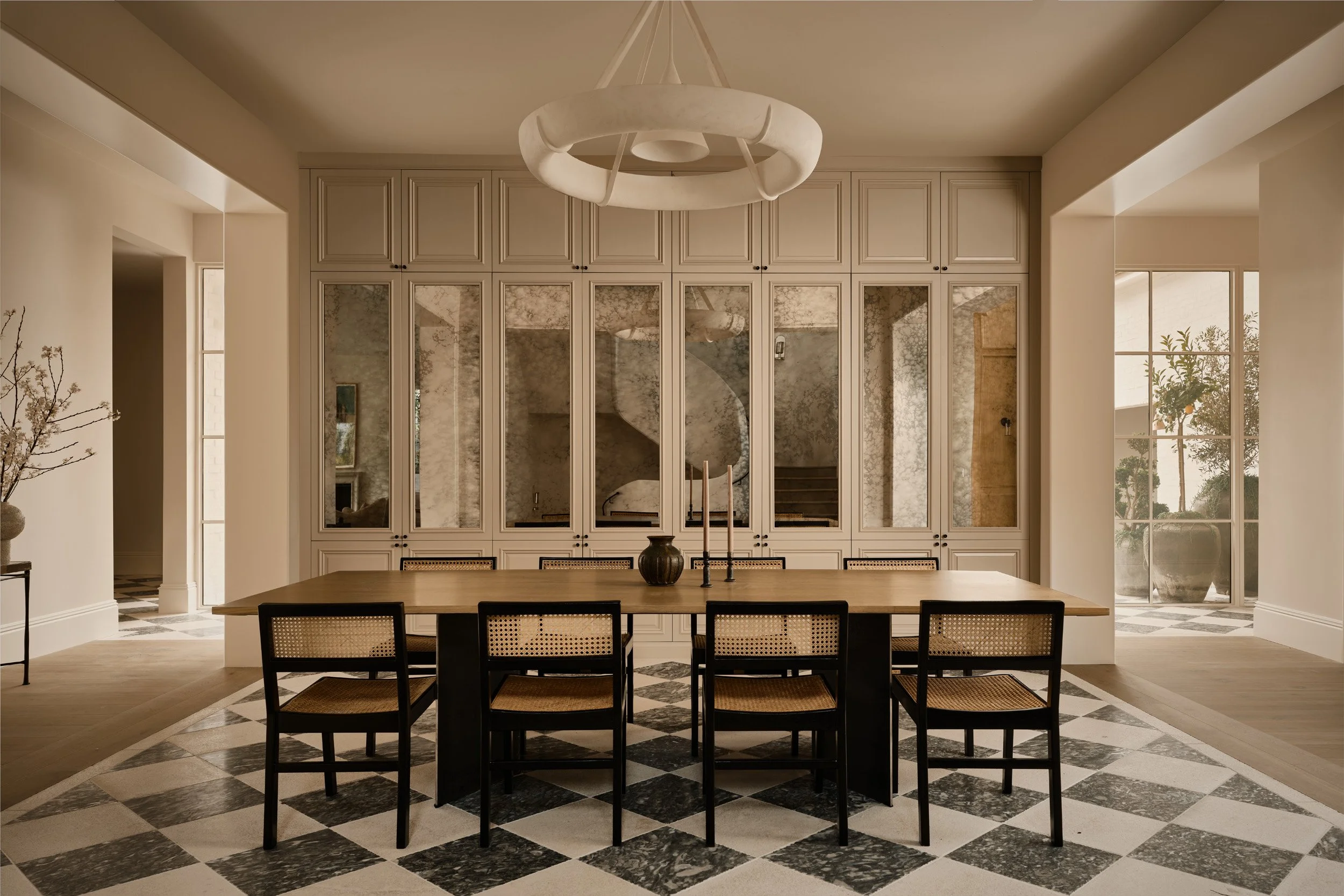 Modern dining room with a wooden table, six chairs, a black vase, and candlesticks. Large mirrors or glass panels behind the table reflect the staircase and walls. Checkerboard marble floor, cream-colored walls, and a unique circular chandelier hanging from the ceiling. Large windows allow natural light, showing outdoor plants.