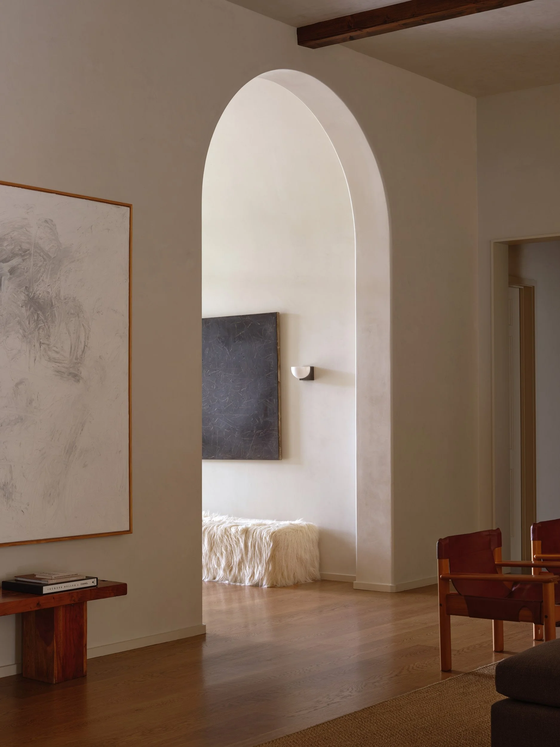 Interior of a modern living room with a wooden floor, a wooden side table with books, a large wall art piece, and a cozy bench with a furry covering, visible through an arched doorway.