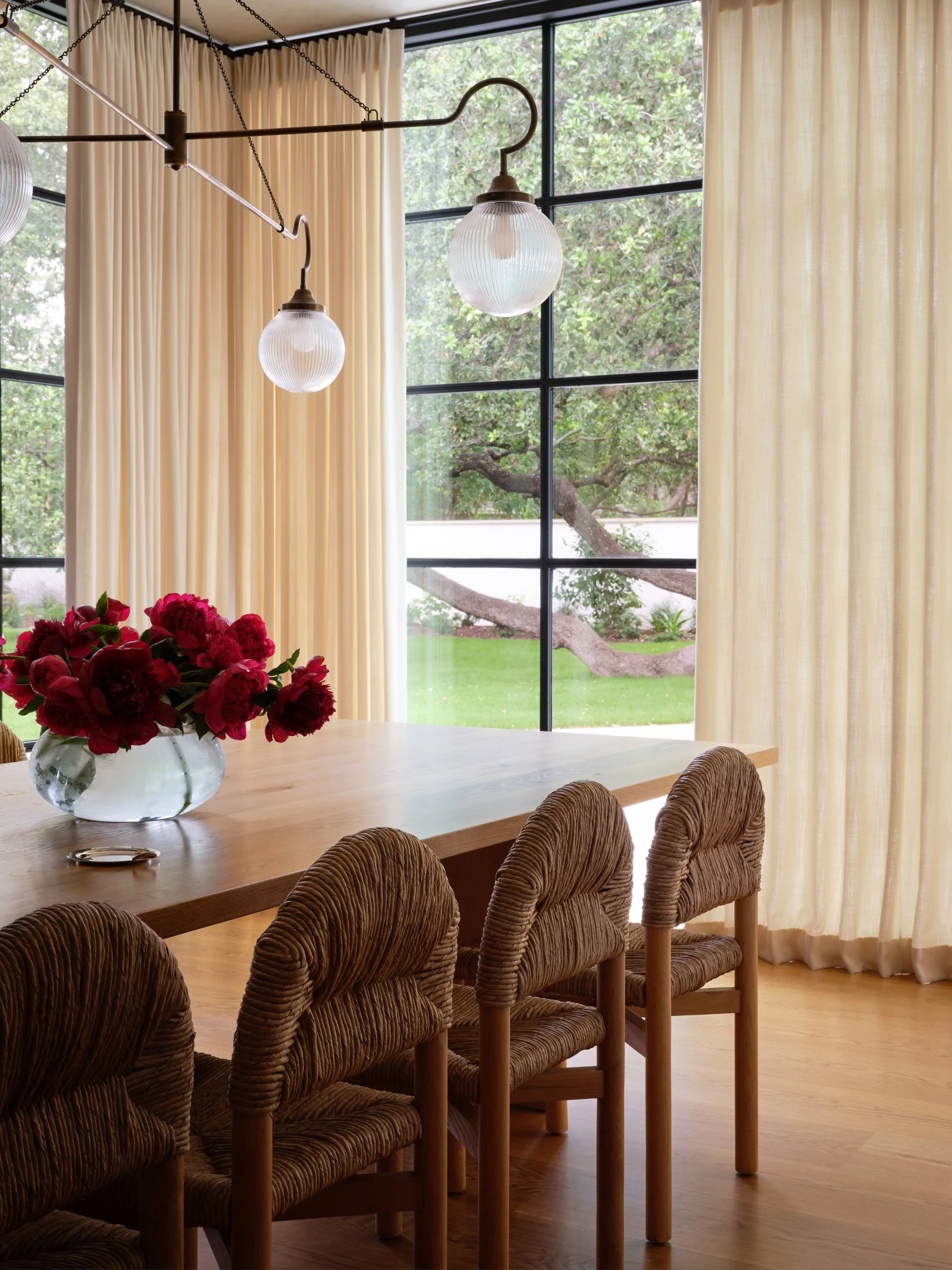 A dining room with a wooden table, four brown textured chairs, a vase of red flowers, large glass windows with beige curtains, and hanging globe pendant lights.