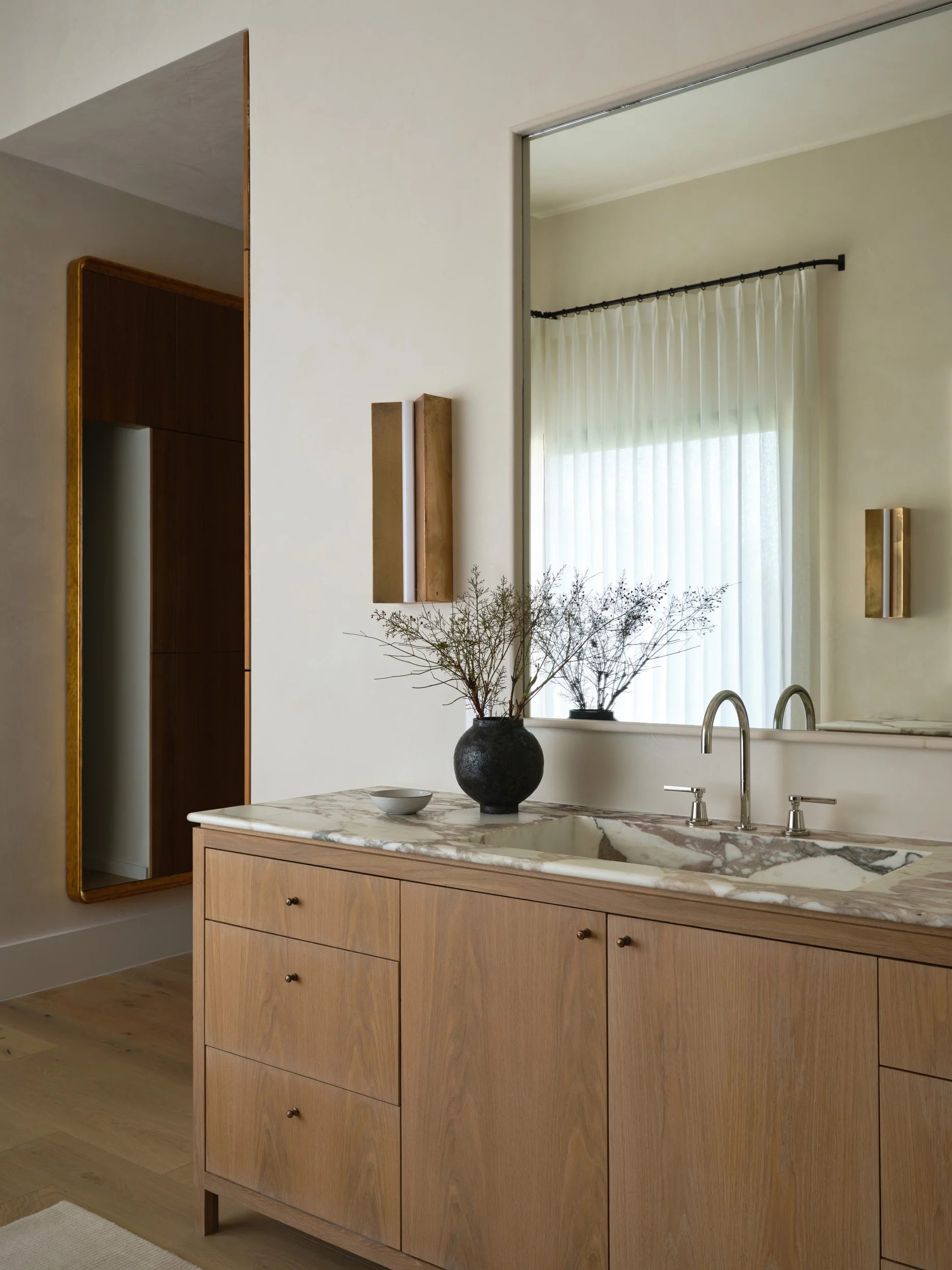 Bathroom vanity with a marble countertop, a black vase with dried branches, a small bowl, a large mirror, and a window with sheer curtains.