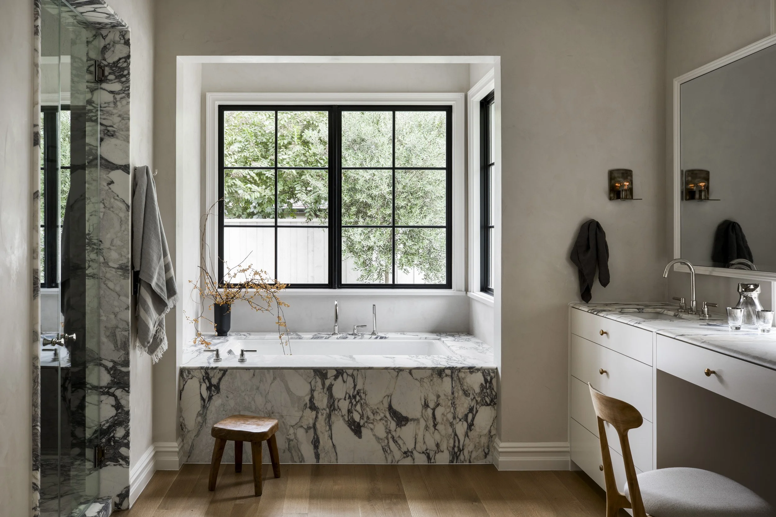 Modern bathroom with marble bathtub and shower, black window frames, wooden stool, and white cabinetry with gold handles.