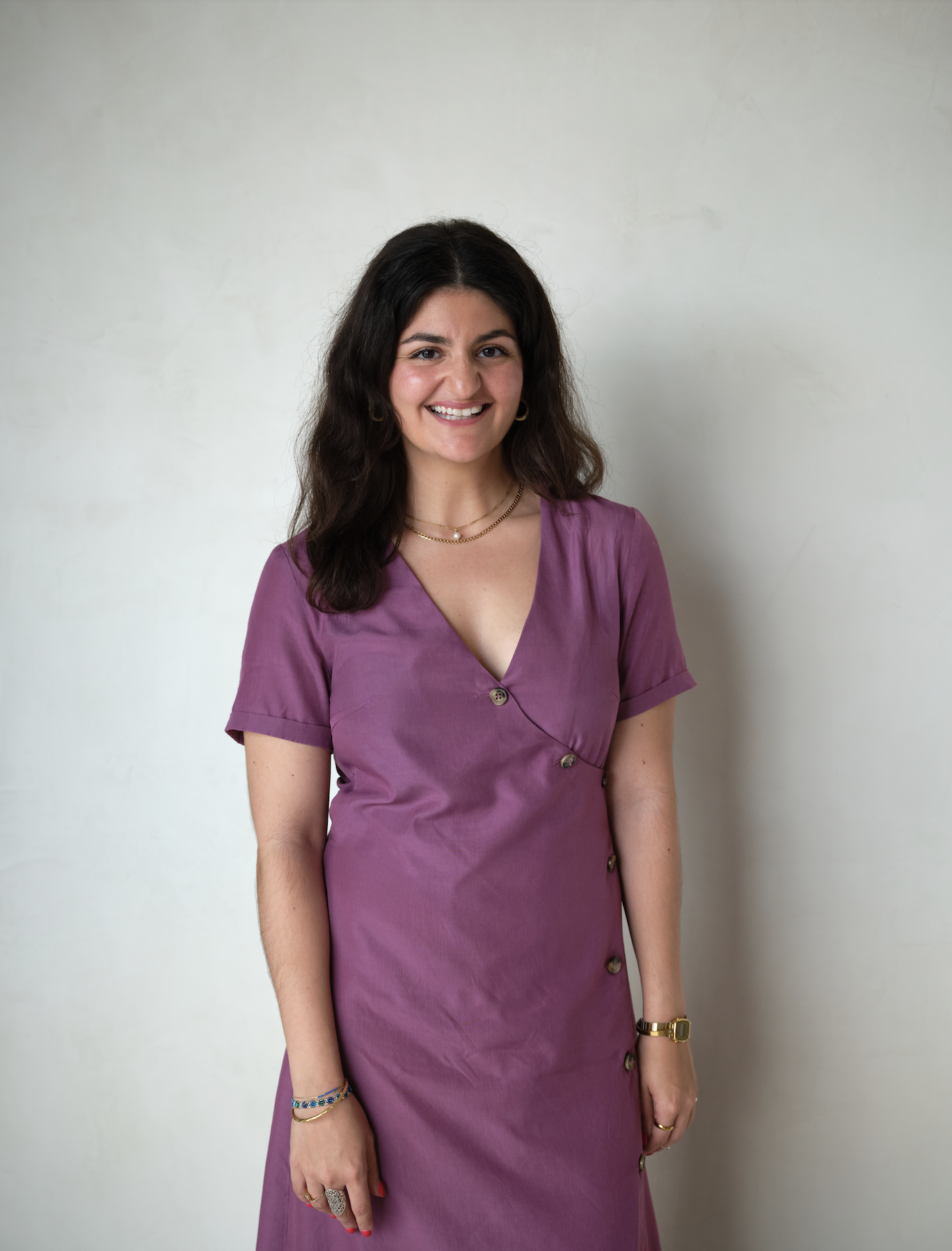 A woman with dark, wavy hair and a smile, wearing a short-sleeved purple dress with buttons, standing against a plain light-colored wall.