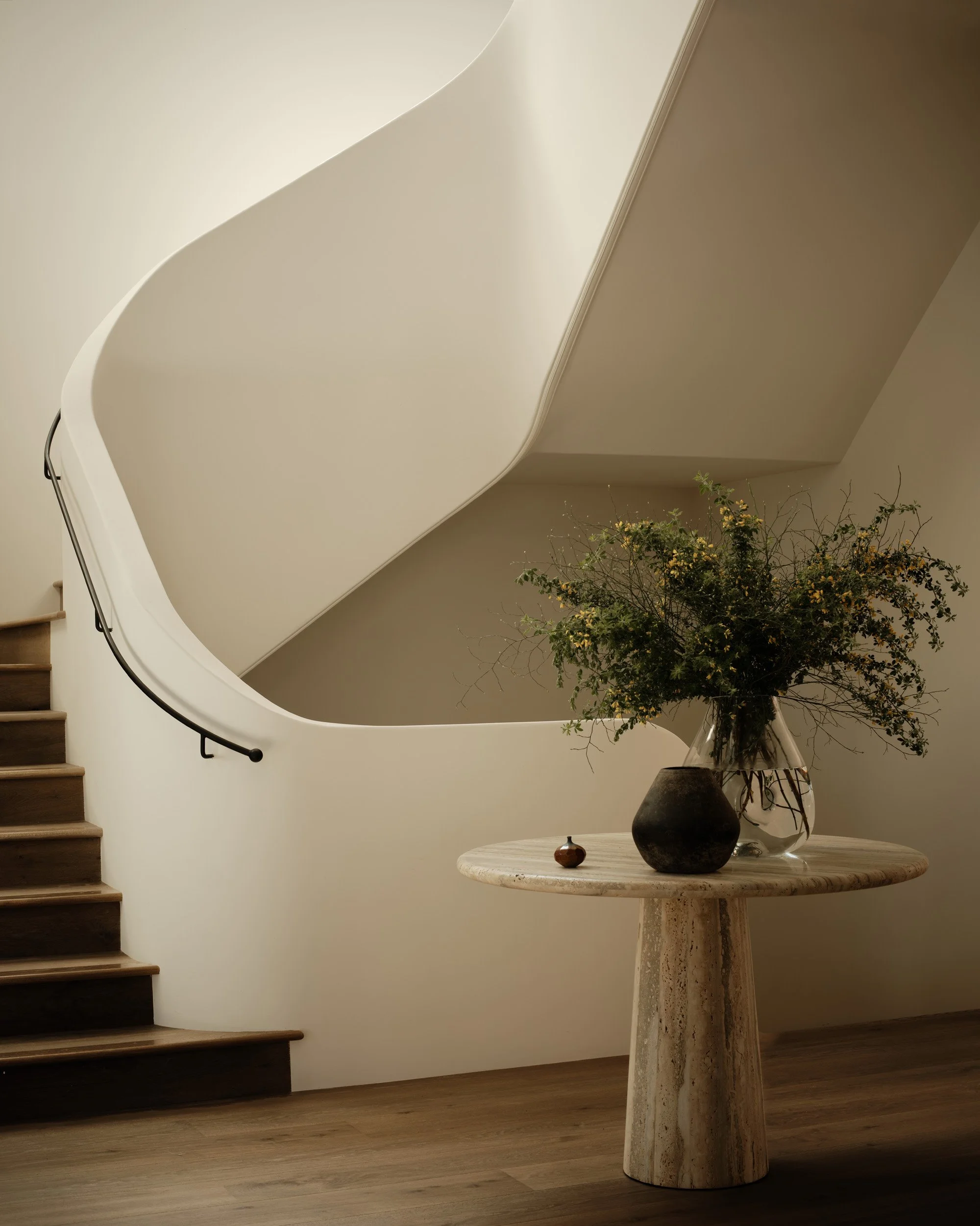 Interior of a modern staircase with wooden steps, a black handrail, a round marble table with a large glass vase of mixed green plants and a smaller dark vase nearby.