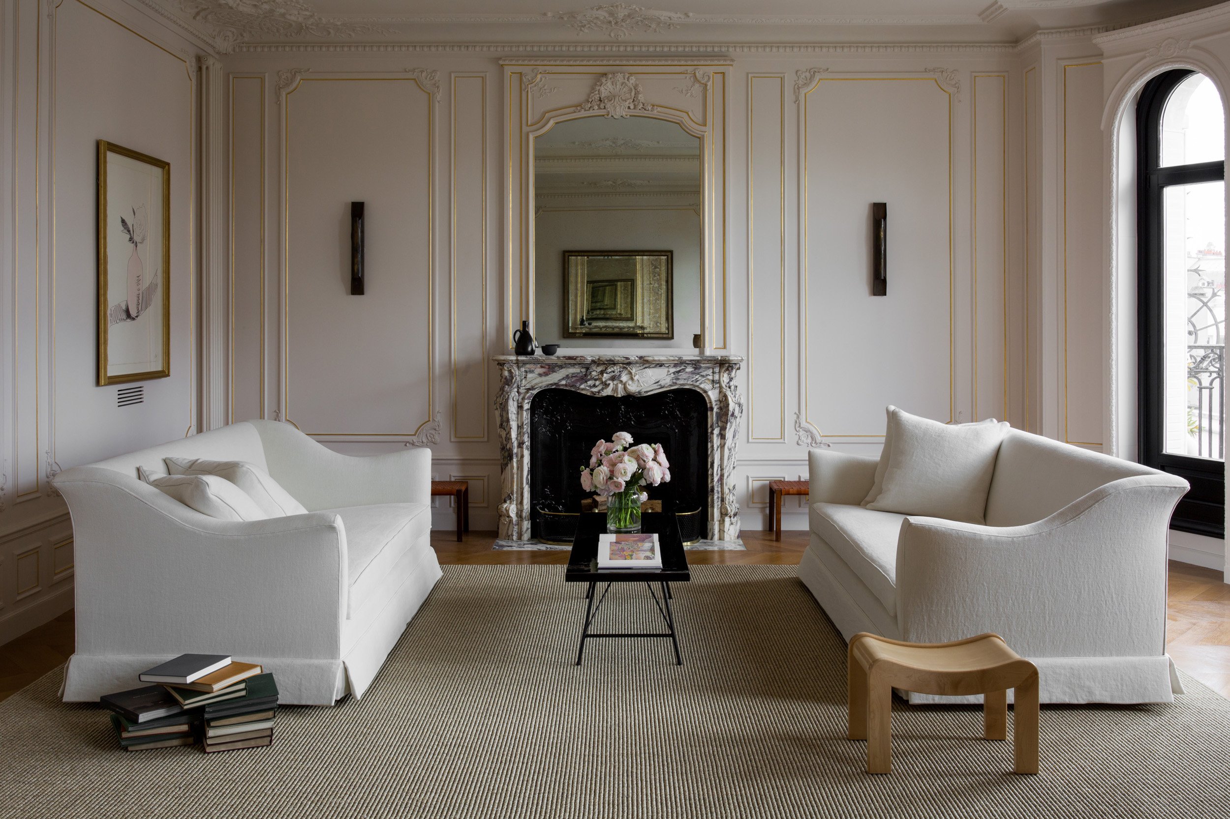 Elegant living room with cream sofas, a black coffee table with a flower arrangement, a marble fireplace, and a large window with a balcony outside.