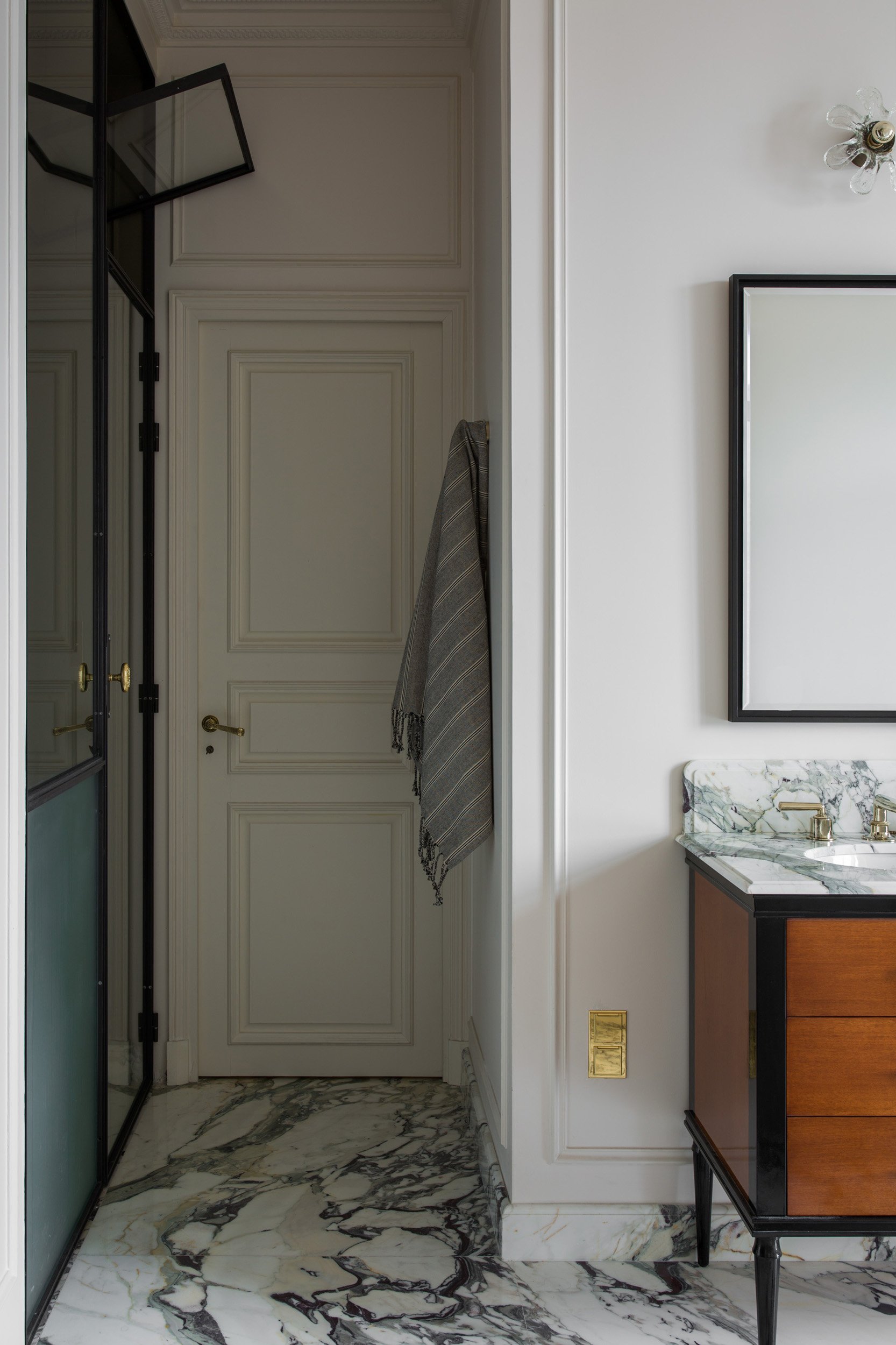 Interior view of a luxurious bathroom with a marble floor, a white paneled door, a towel hanging on the wall, part of a wooden vanity with a marble countertop and a mirror, and a wall sconce light