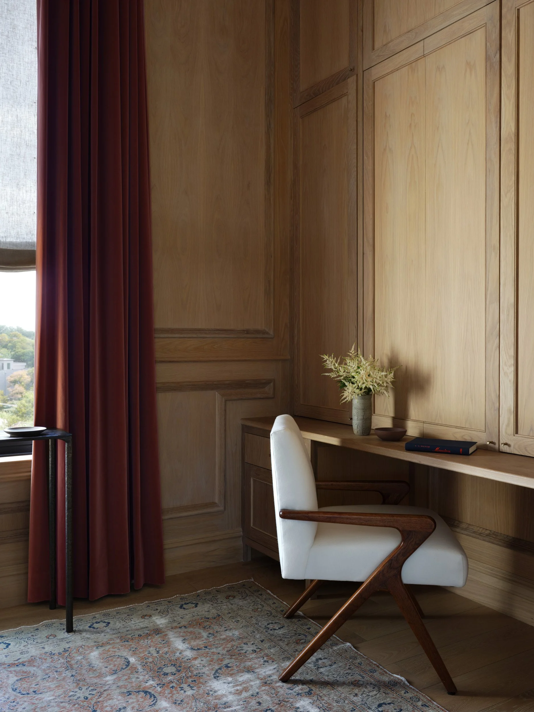 A cozy corner with a wooden desk, a white modern armchair, and a vase of white flowers, with a window and red curtains in the background.