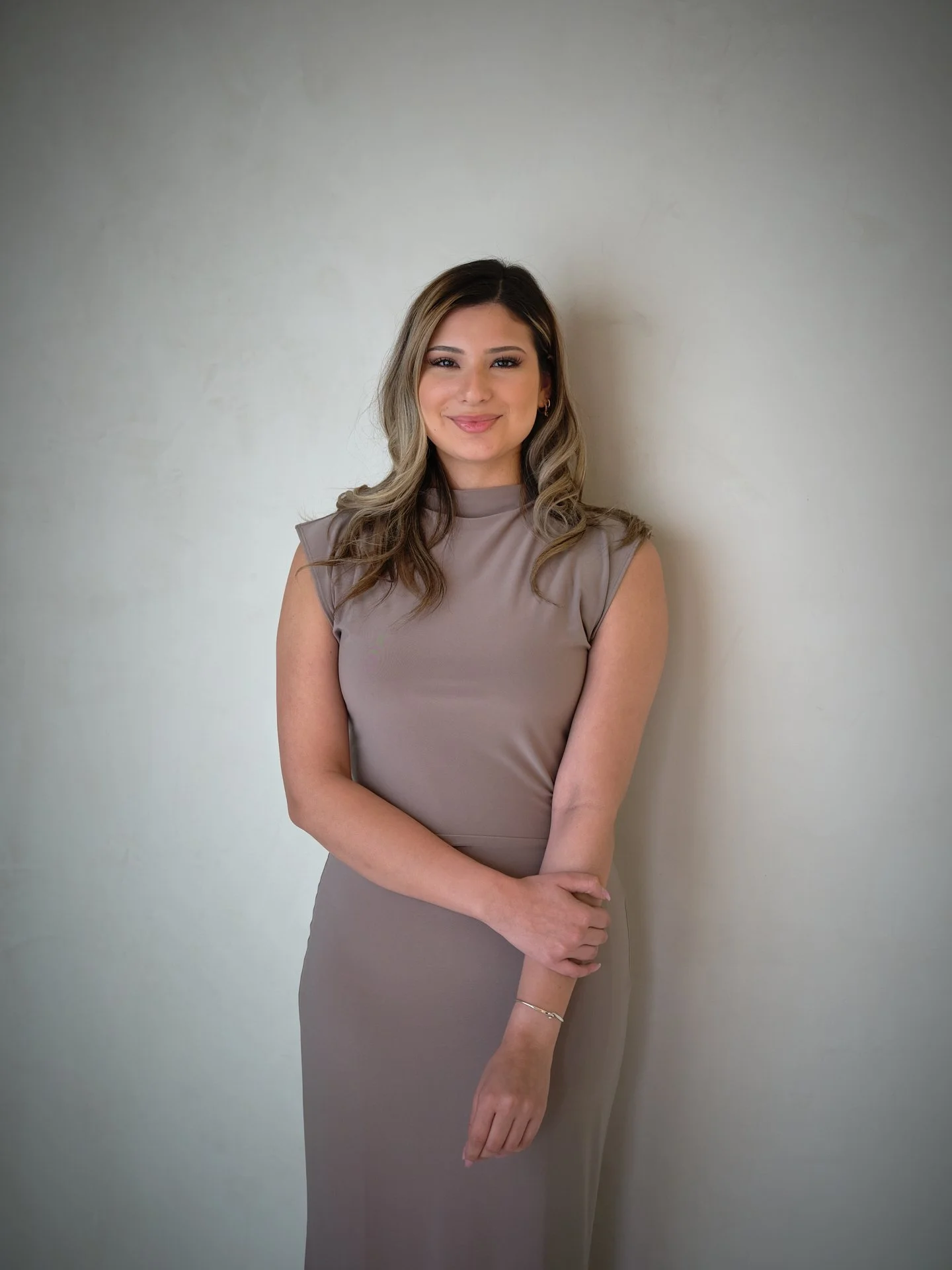A woman with long, wavy blonde hair and wearing a taupe dress, standing against a plain light-colored wall, smiling softly at the camera.