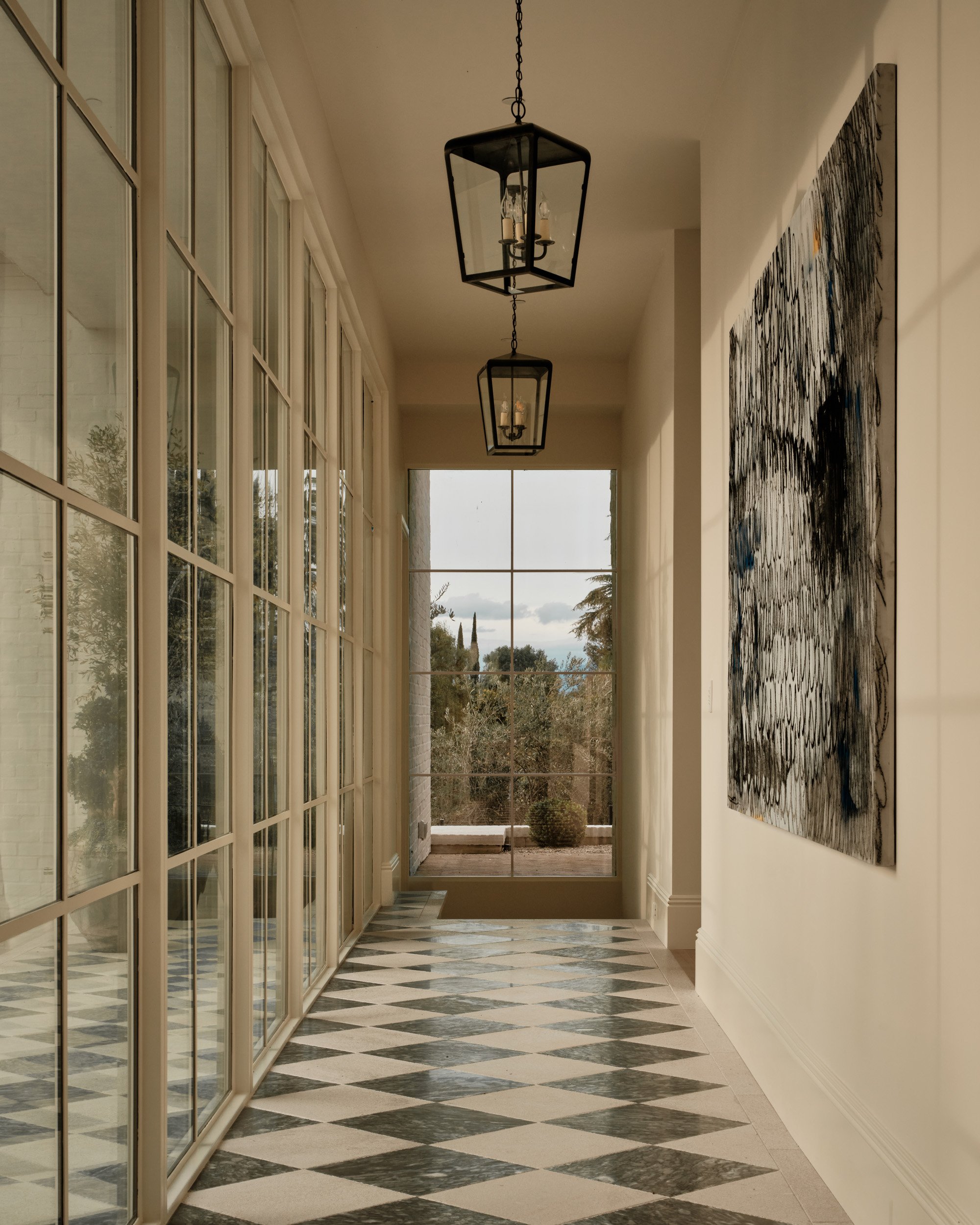 A hallway with large glass paneled sliding doors on the left, black metal lantern pendant lights hanging from the ceiling, a large abstract black and white painting on the right wall, and a window at the end showing trees and a cloudy sky outside.