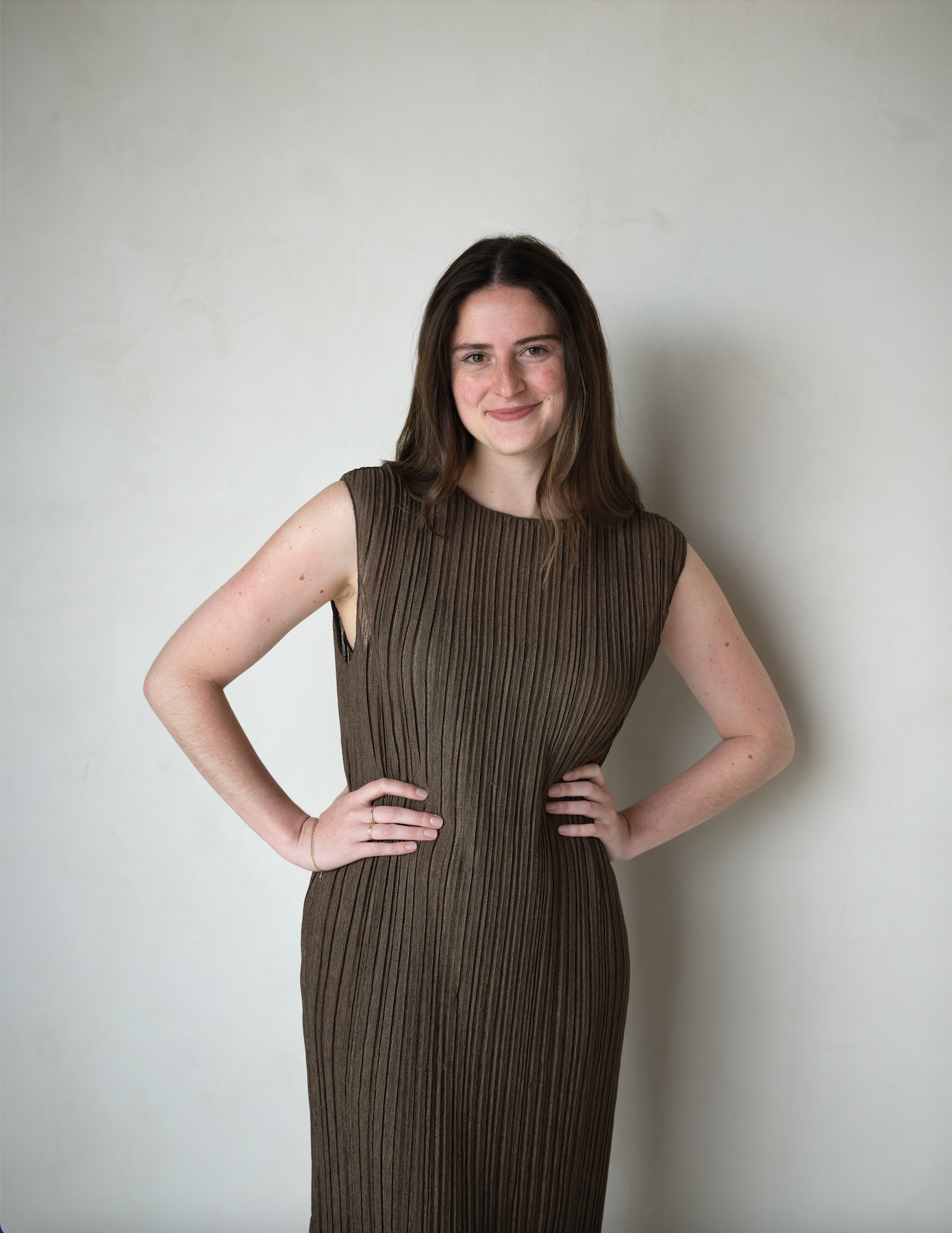 A woman with shoulder-length brown hair wearing a sleeveless brown pleated dress, standing with her hands on her hips against a light plain wall, smiling at the camera.