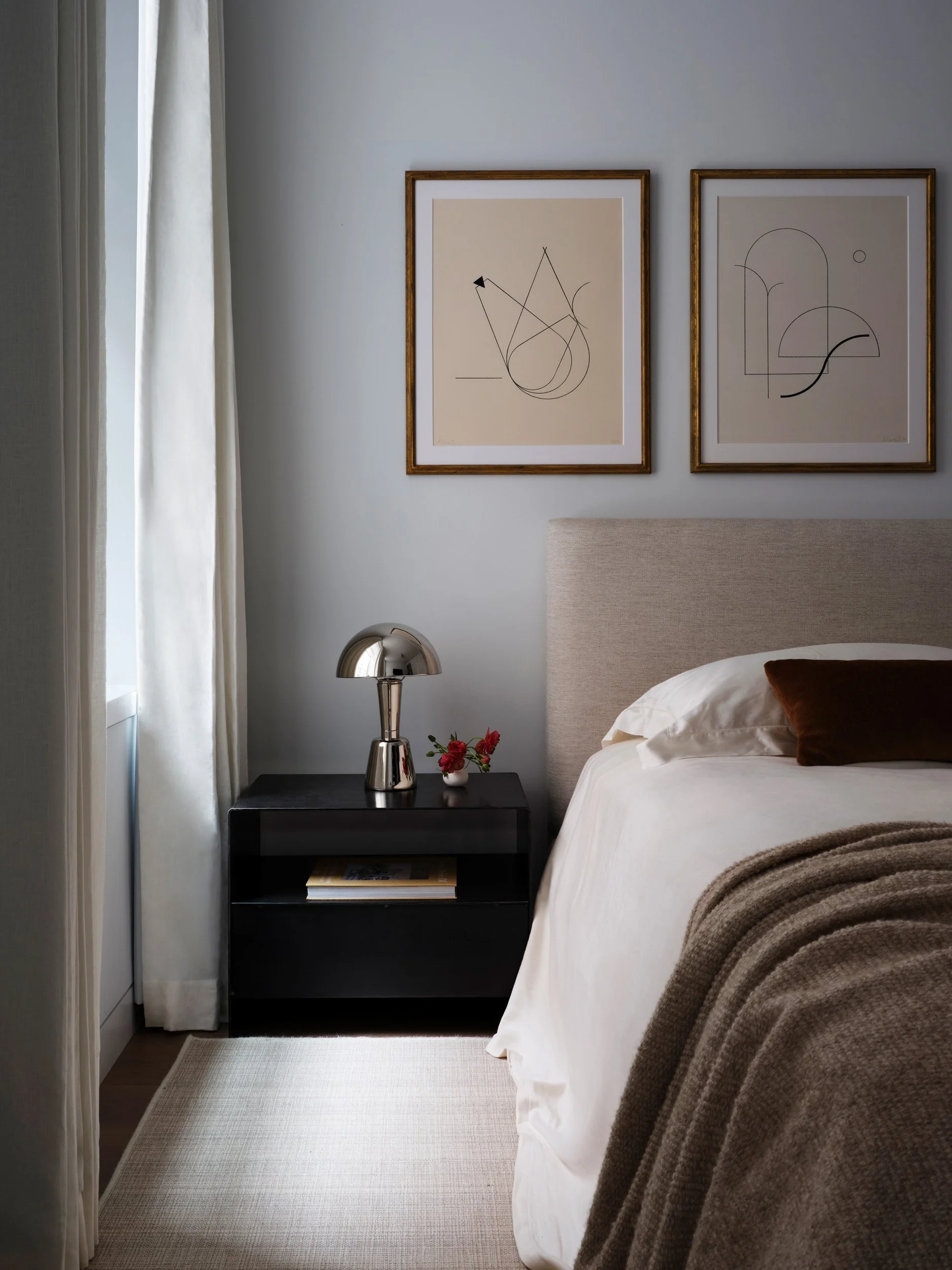 A cozy bedroom corner with a bed, beige headboard, white bedding, and a brown pillow. Beside the bed is a black nightstand with a silver modern lamp, a small vase of red flowers, and a book. Above the bed are two framed abstract line art sketches on a white wall, with a light-colored curtain on the window to the left.