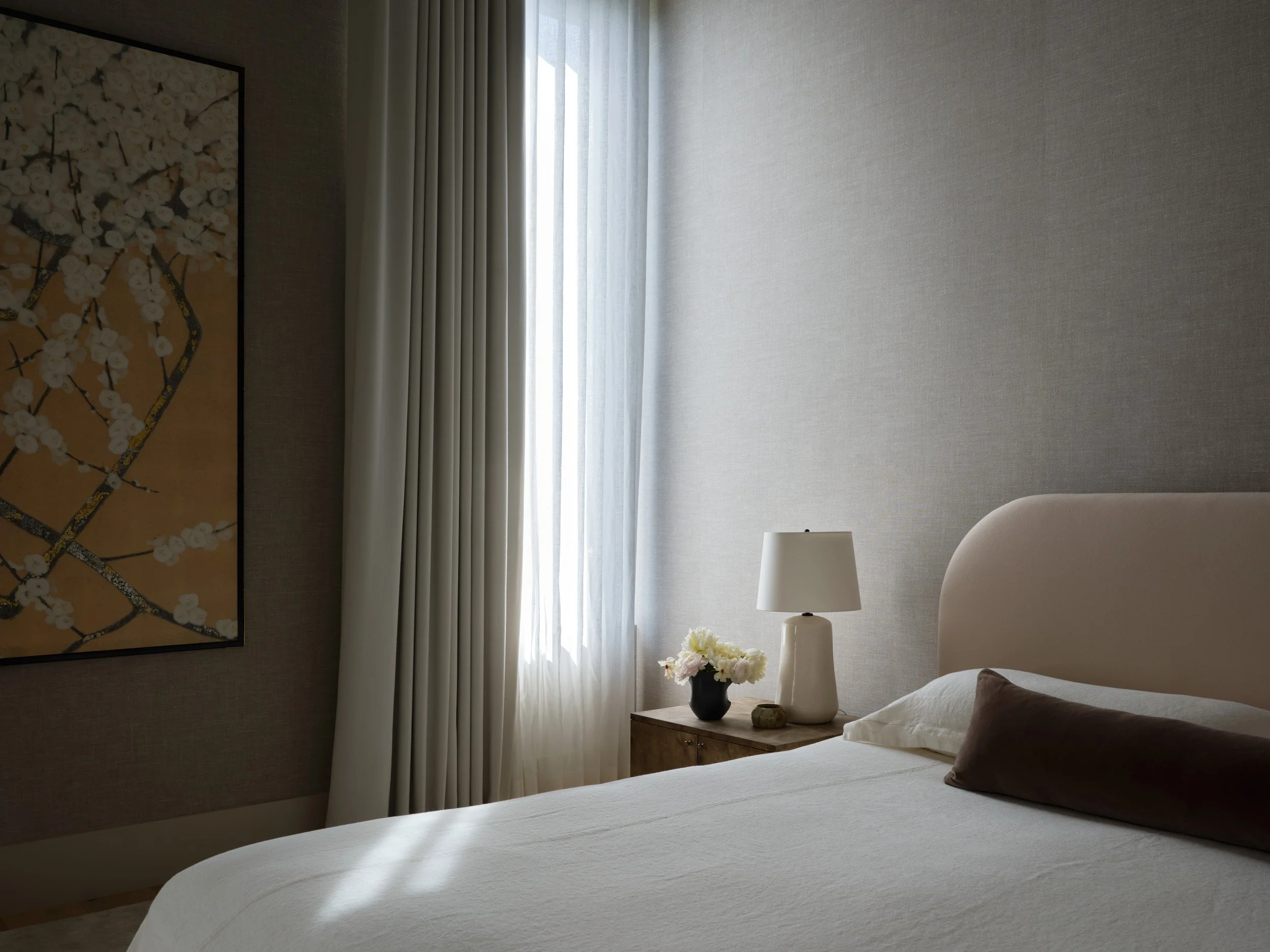 A bedroom with a bed, pillow, a bedside table with a lamp, a flower vase, and a piece of artwork on the wall, illuminated by daylight through window curtains.