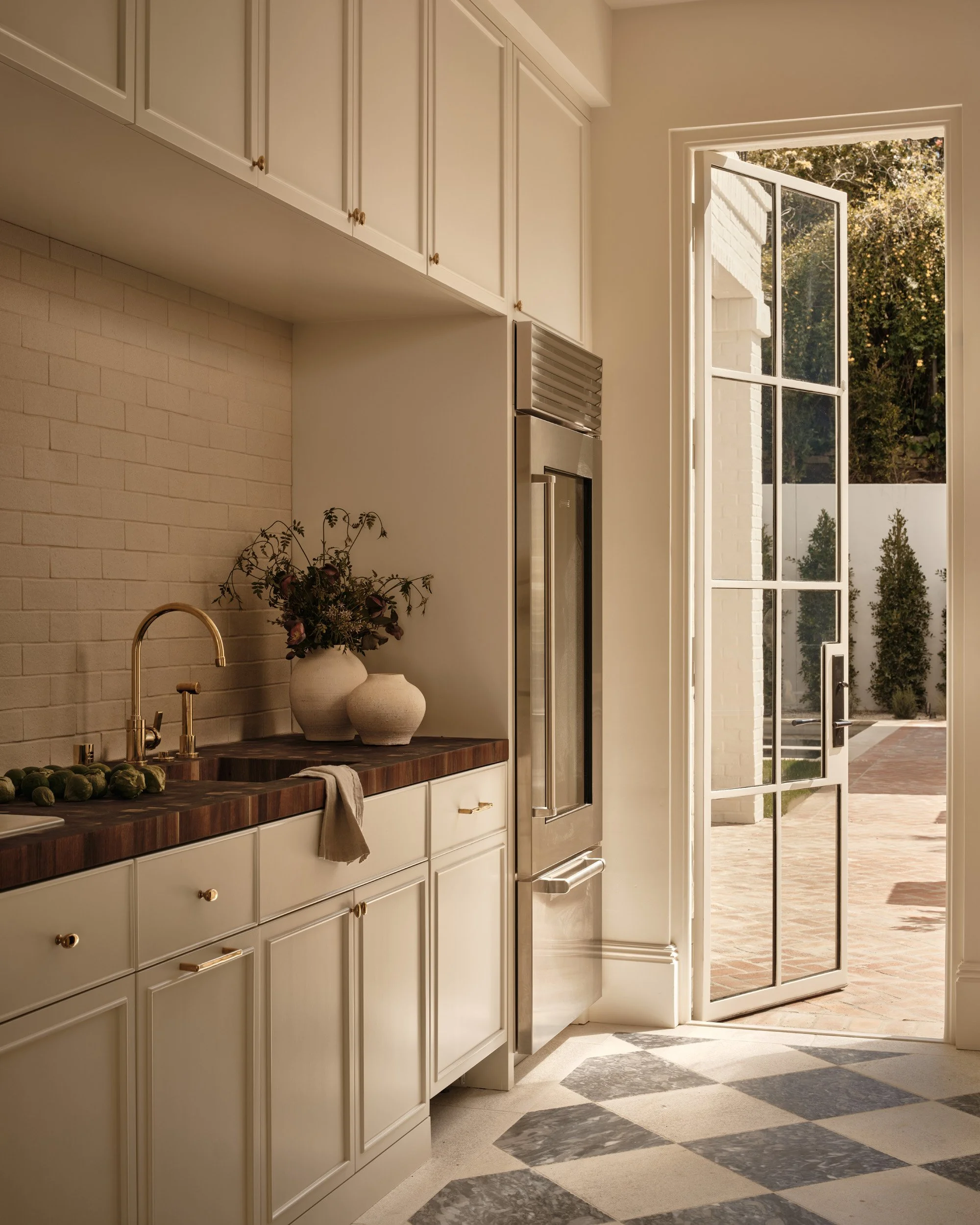 Kitchen with cream-colored cabinetry, a wooden countertop, a gold faucet, decorative vases with flowers, and an open door leading outside to a patio with brick paving and greenery.
