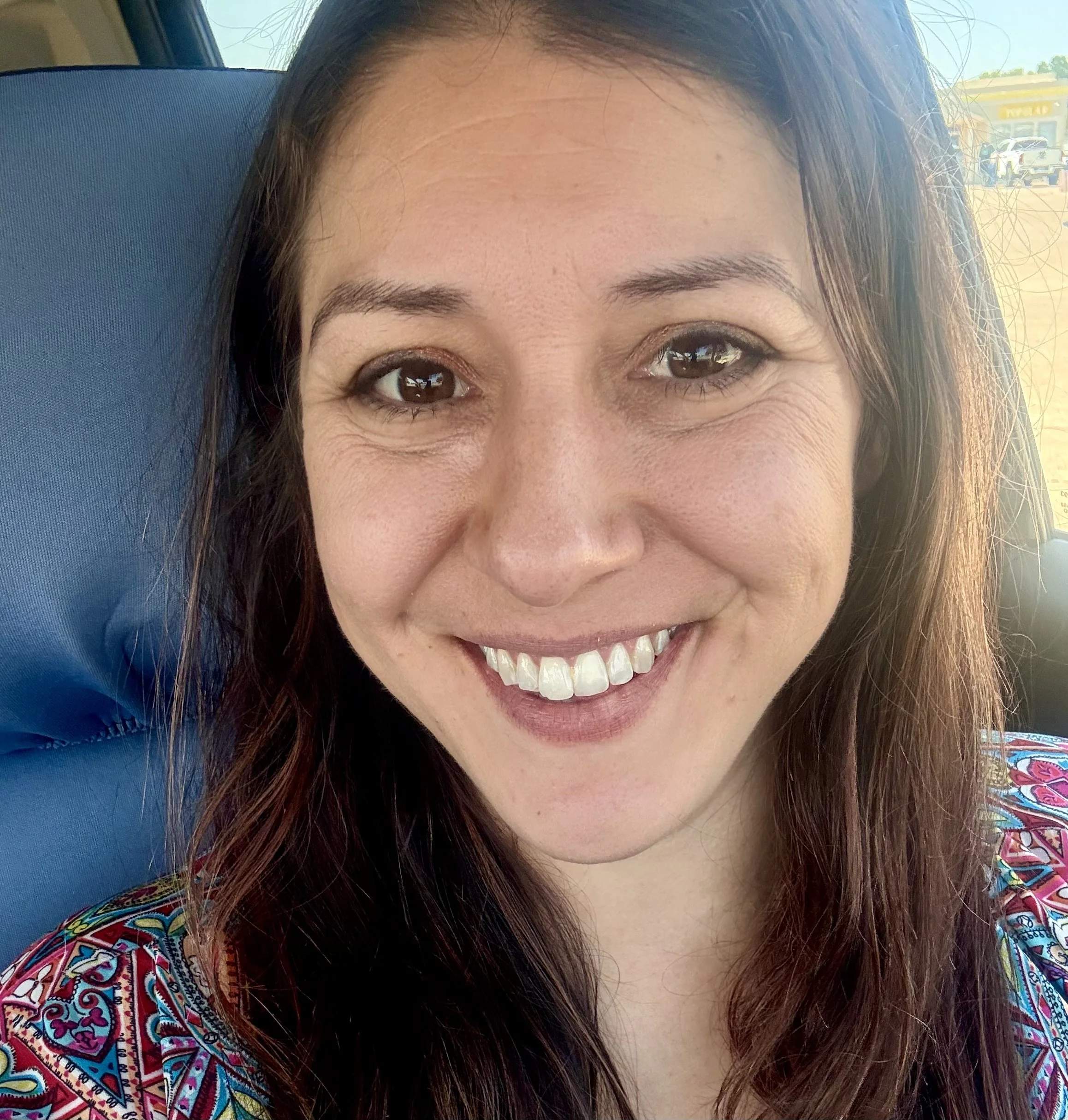 Close-up of a smiling woman with long dark hair, sitting on a blue seat inside a vehicle, with sunlight and a parking lot visible in the background.