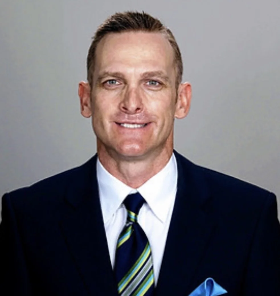A man with short, light brown hair, blue eyes, wearing a navy suit, white shirt, striped tie, and a blue pocket square, smiling against a plain gray background.